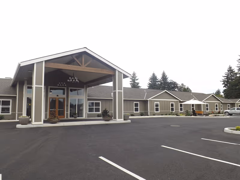 Exterior view of Cherrywood Memory Care facility showing a single-story building with a covered entrance supported by columns, several windows, a parking lot in front, and trees in the background under an overcast sky.