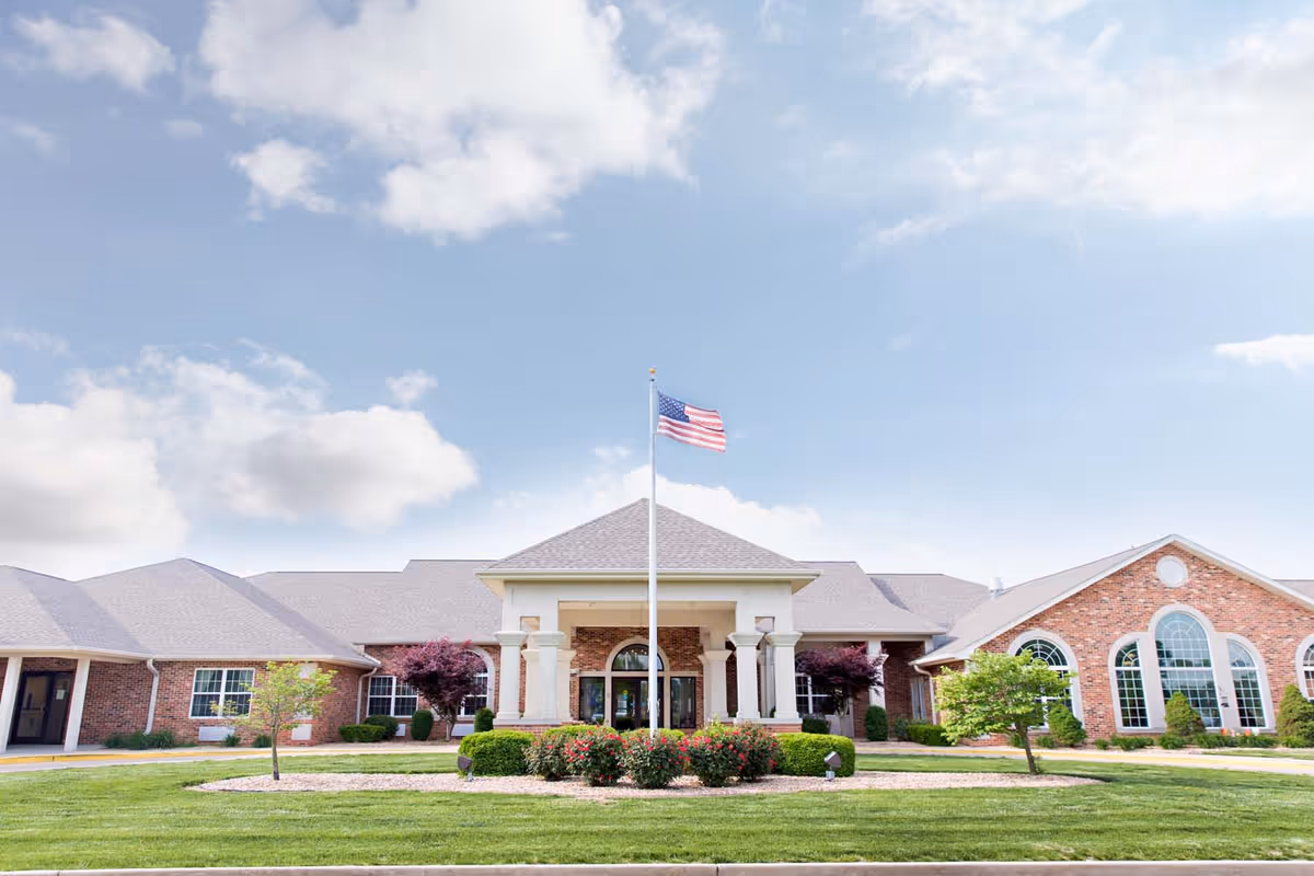 Front entrance of a brick retirement facility with an American flag on a flagpole and landscaped lawn.