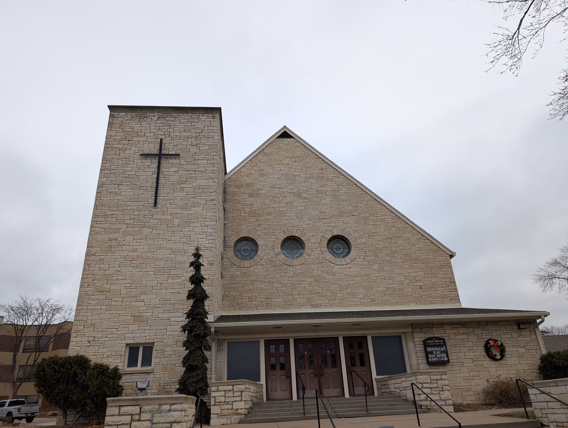 Front exterior view of a stone church building with a large cross mounted on the left tower, three circular stained glass windows on the main facade, and a wreath hanging on the right side near the entrance doors.