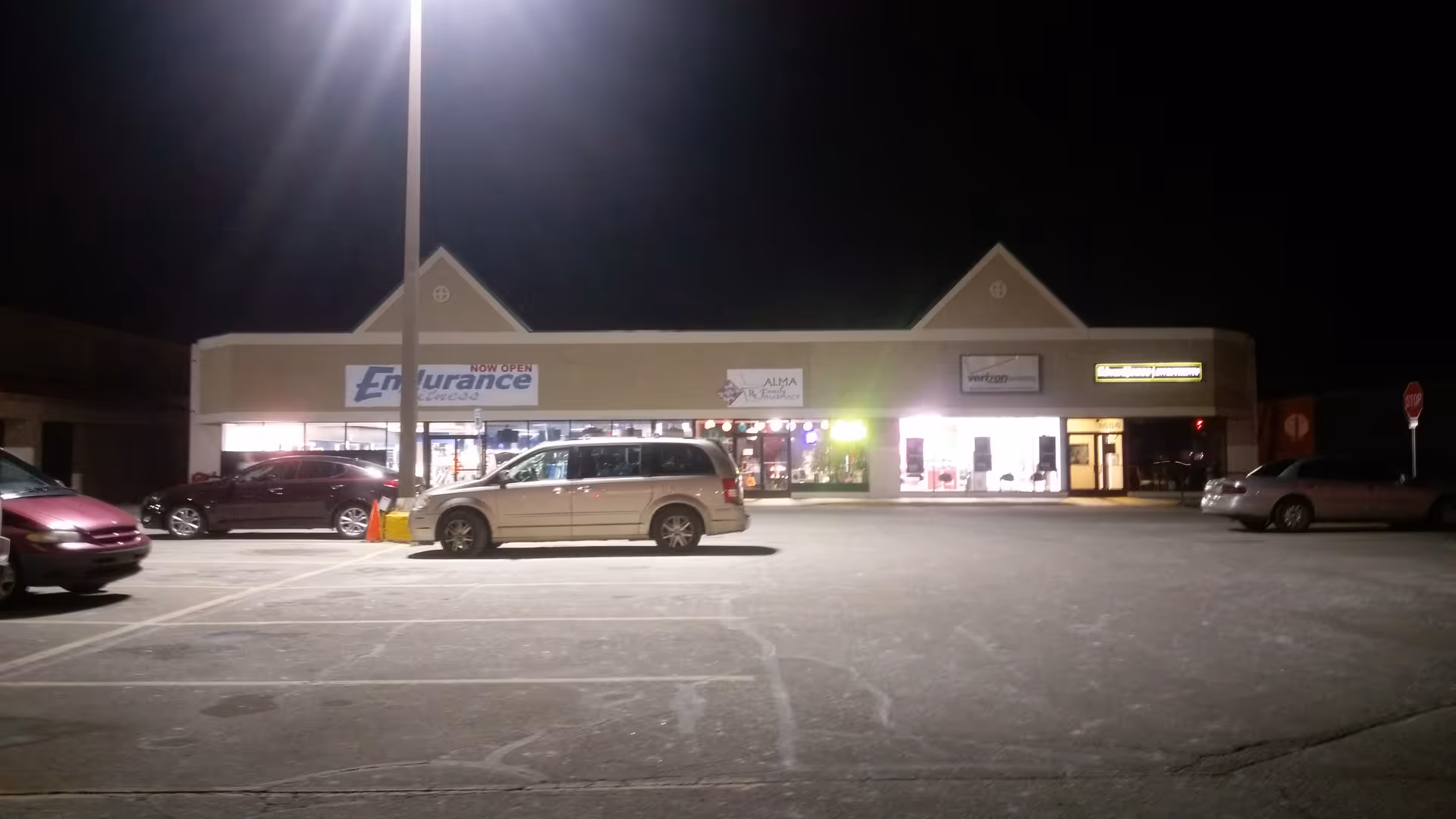 Nighttime exterior of a strip mall with lit storefronts and parked cars.