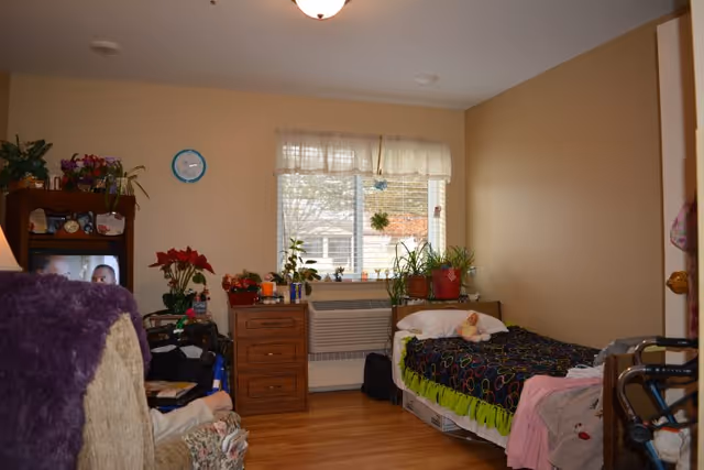 A small, cozy bedroom with beige walls and wooden flooring. There is a single bed with colorful bedding and a pillow, positioned against the right wall. Next to the bed is a wooden nightstand with several potted plants on top. A window with sheer curtains lets in natural light. On the left side of the room, there is a TV on a stand with more plants and personal items around it. A walker is partially visible near the bed, indicating accessibility accommodations.