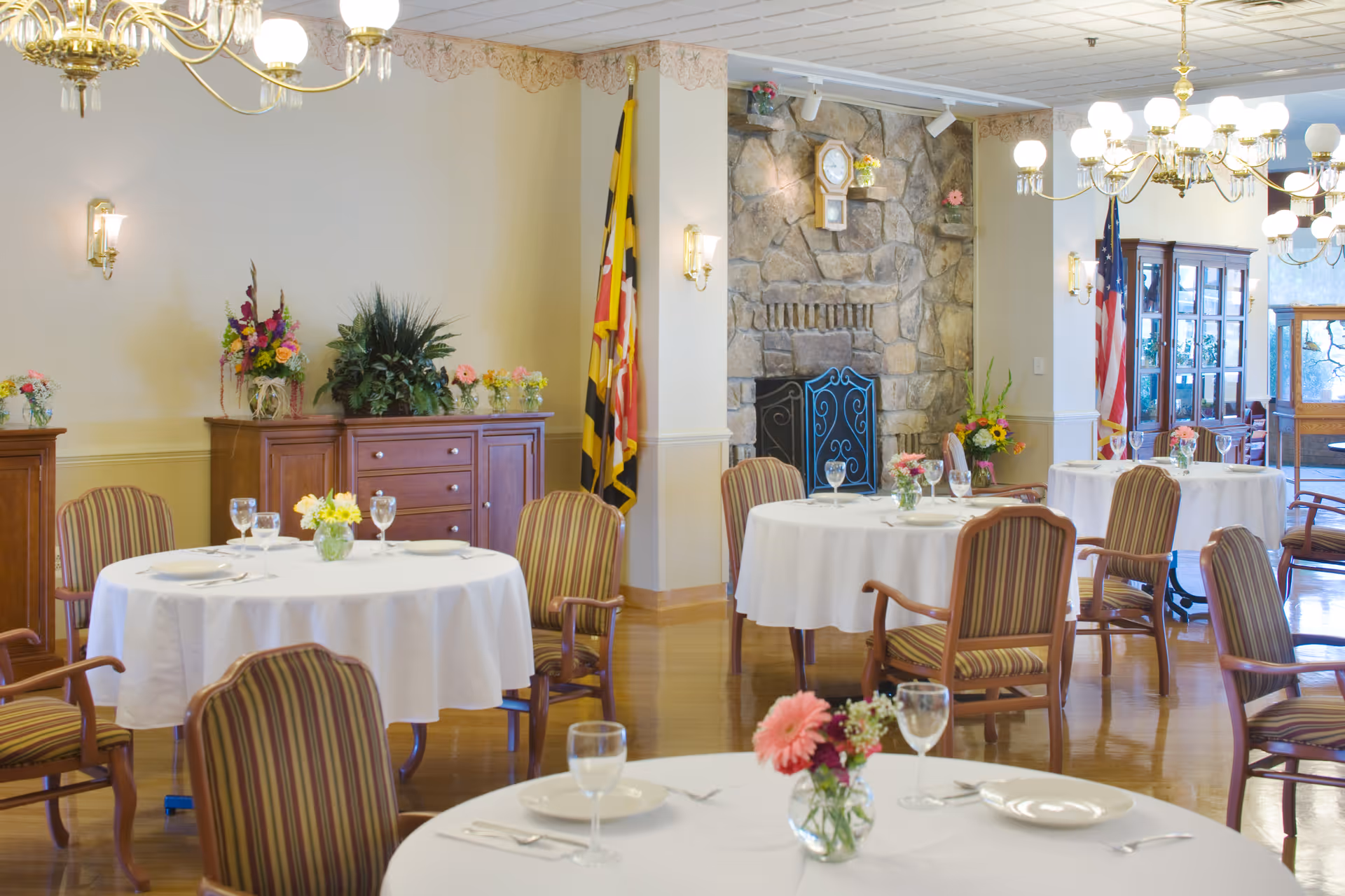 A bright dining room with round tables covered in white tablecloths, each set with plates, glasses, and silverware. The room features striped upholstered chairs, floral centerpieces on each table, a stone fireplace with a clock above it, and flags in the corner. Wooden cabinets with plants and flowers are against the walls, and chandeliers hang from the ceiling.