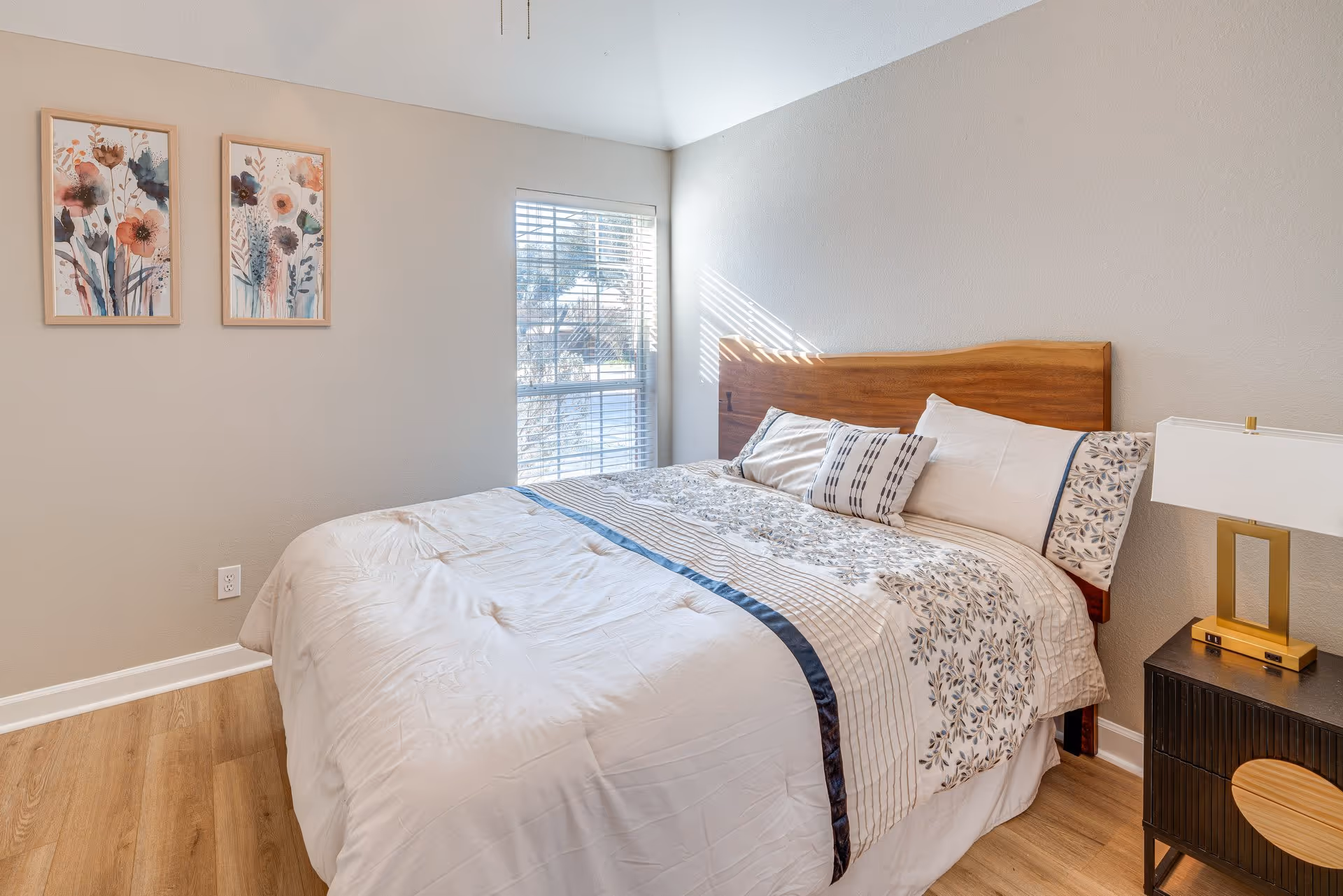 A neatly made bedroom with a wooden headboard, bedding, nightstand, and sunlit window.