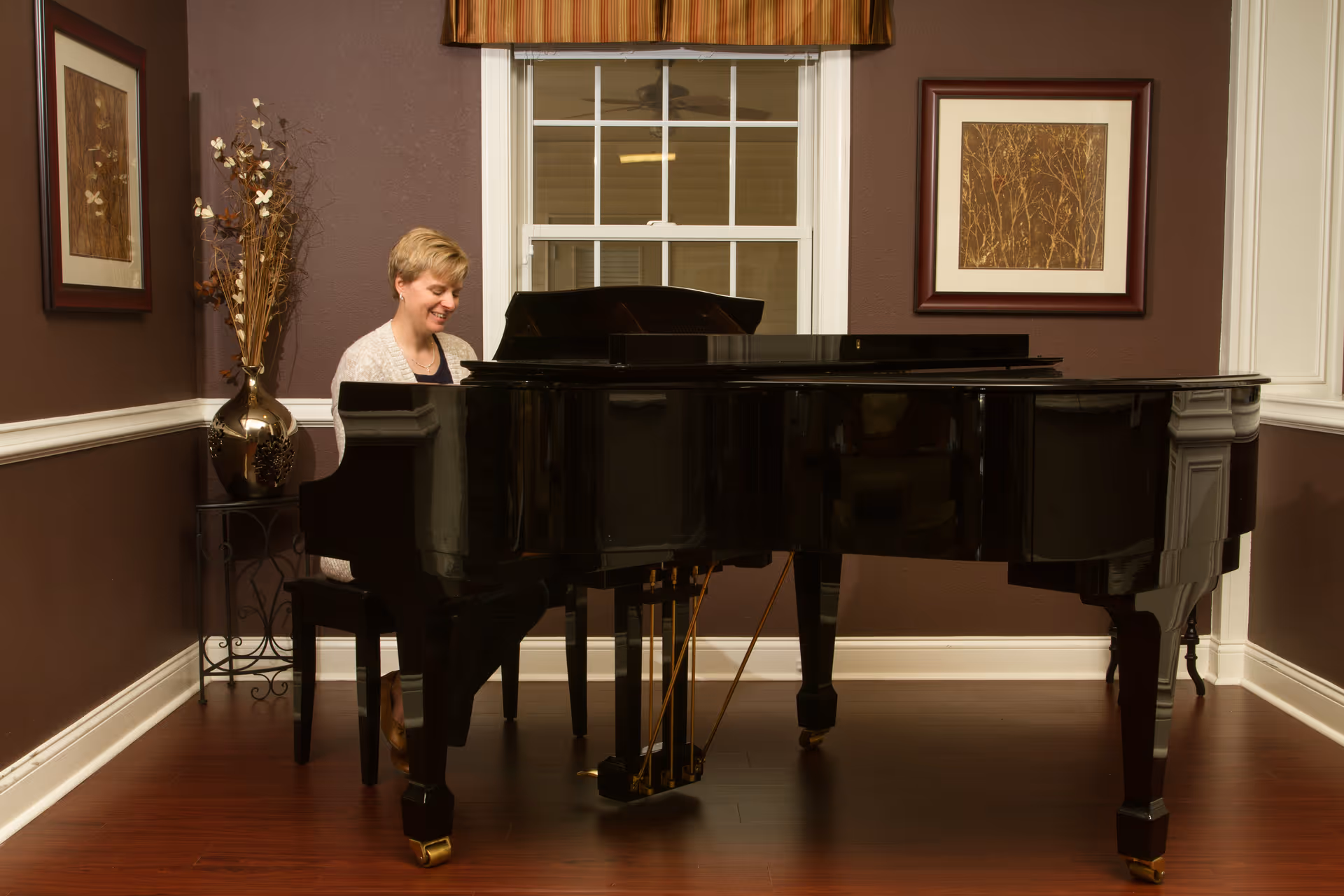 A woman sitting and playing a black grand piano in a warmly decorated room with brown walls, two framed pictures, a window with a striped valance, and a decorative vase with dried flowers on a small table.