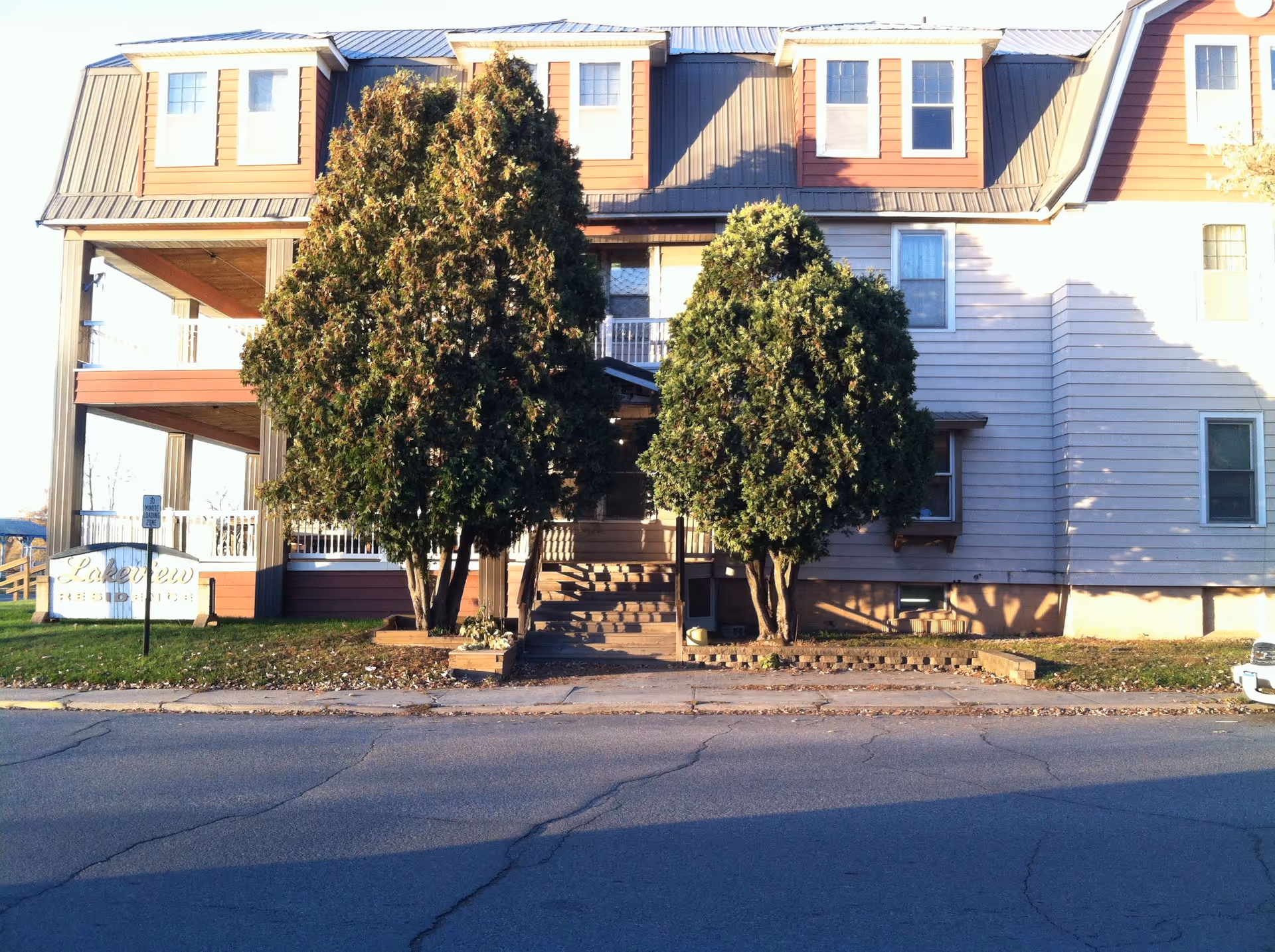 Exterior view of a multi-story residential building with a metal roof and several windows. Two large evergreen trees are in front of the entrance, which has stairs leading up to a covered porch. A sign near the left side reads 'Lakeview Residence'. The building is situated next to a paved road.
