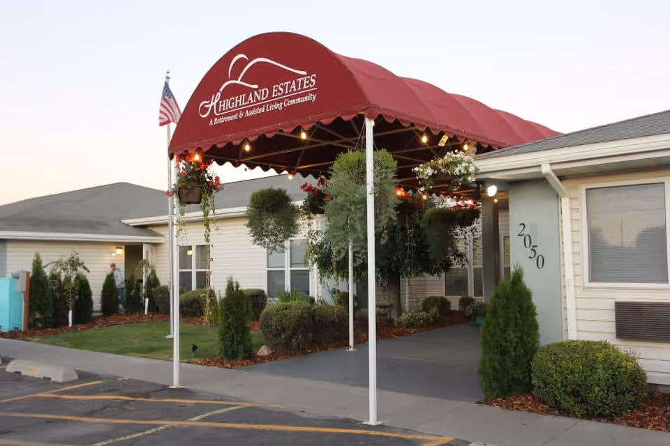 Entrance of Highland Estates Assisted Living & Memory Care facility with a red canopy displaying the facility name. The building is single-story with beige siding, surrounded by small bushes and trees. An American flag is visible near the entrance.