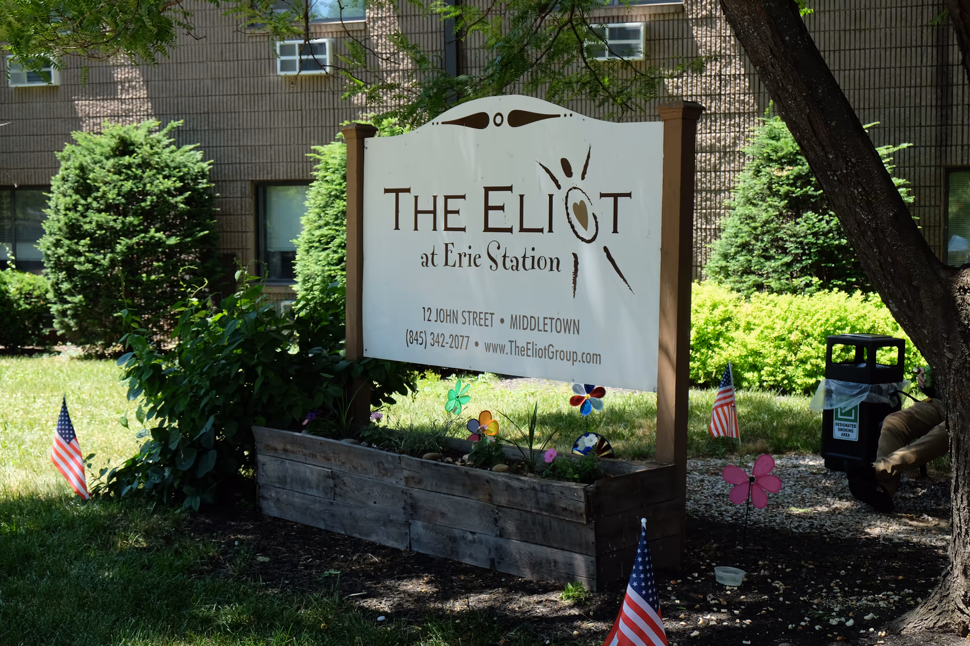 A wooden sign reading "The Eliot at Erie Station" stands in a planted bed in front of shrubs and a brick building.