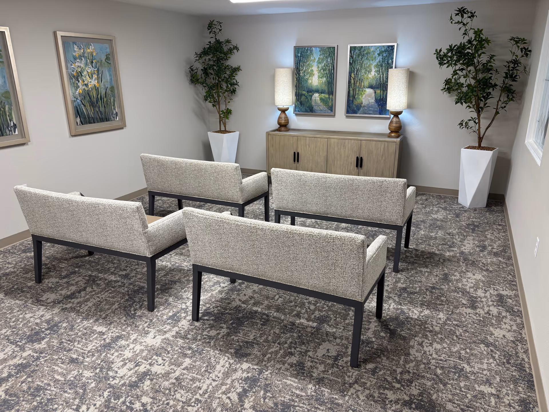 Small waiting room with beige chairs, a cabinet, two lamps, and framed nature art.