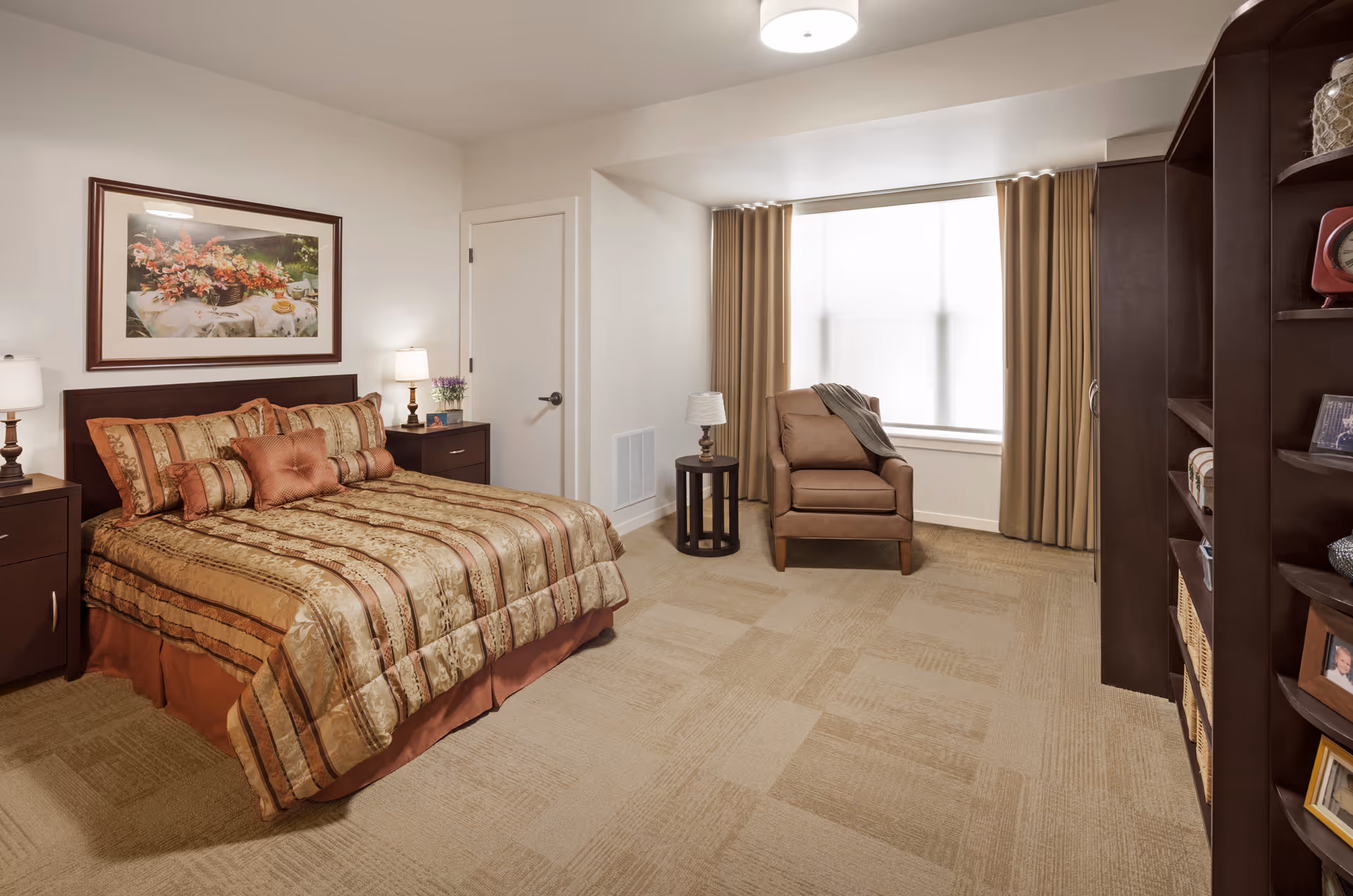 A well-lit bedroom with a large bed covered in a patterned brown and gold bedspread with multiple pillows. On either side of the bed are dark wood nightstands with lamps. A framed floral painting hangs above the bed. To the right, there is a comfortable armchair next to a small round table with a lamp, positioned in front of a large window with beige curtains. On the far right, there is a dark wood shelving unit with various decorative items and baskets.