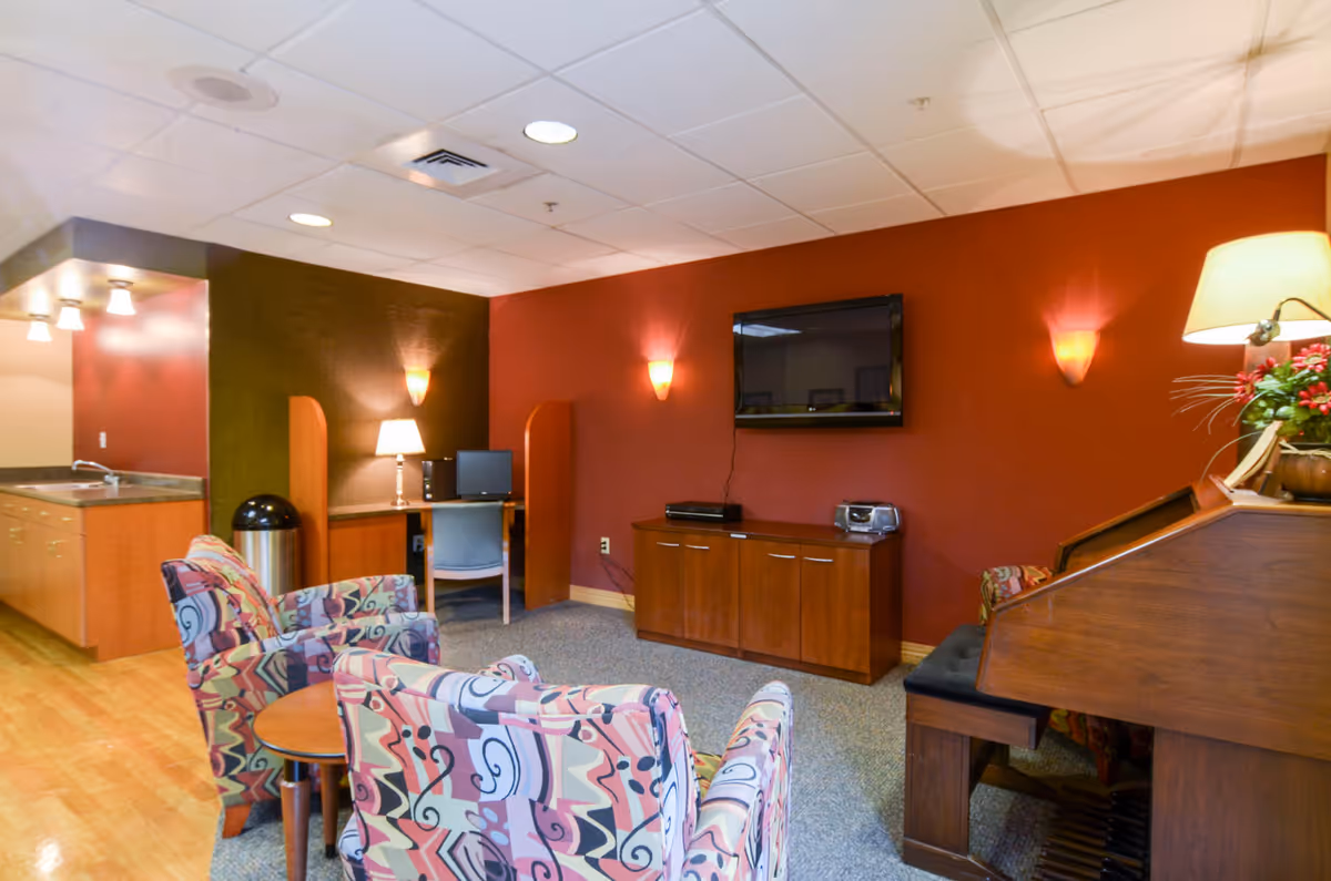 Cozy senior living lounge with patterned chairs, a piano, TV, and a kitchenette.