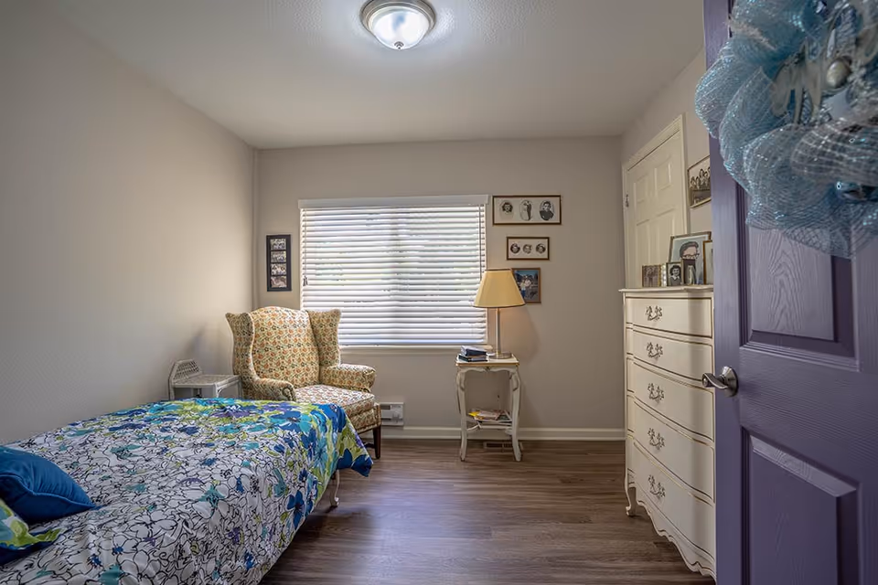 Cozy resident bedroom featuring bed, floral chair, dresser, window, and purple doorway.