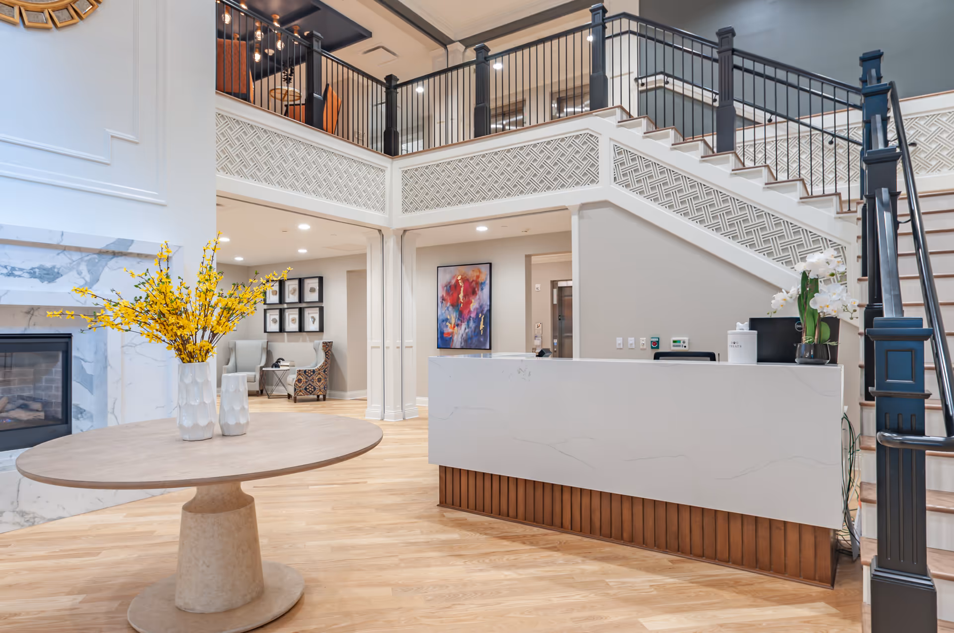 A bright and spacious interior lobby area featuring a modern reception desk with a white marble-like surface and wooden base. To the left, there is a round table with two vases holding yellow flowers, and a fireplace with a marble surround. A staircase with black railings leads to an upper level with a balcony. The walls are light-colored with decorative molding, and there is a colorful abstract painting on the wall behind the reception desk. Comfortable seating is visible in the background.