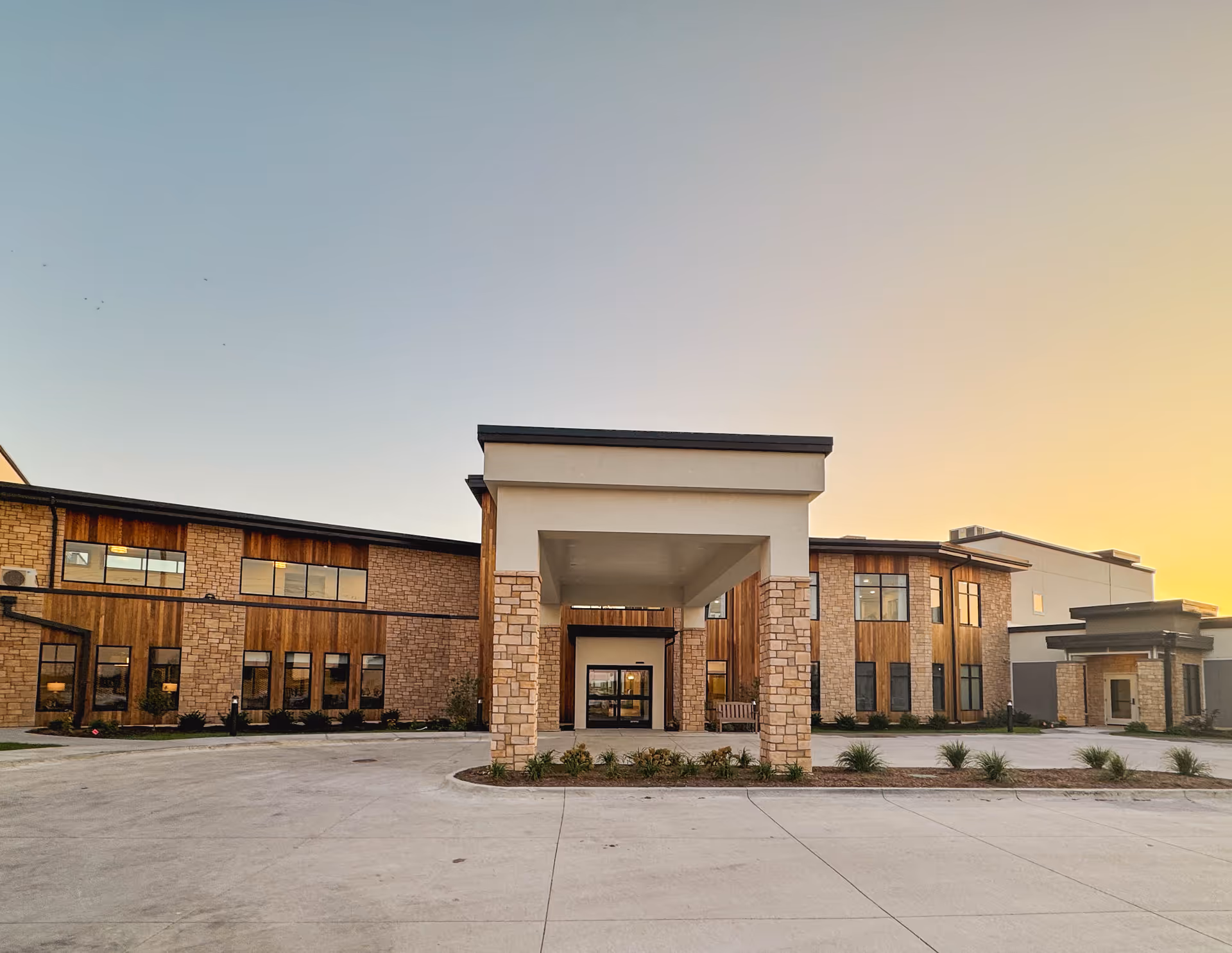 Exterior view of a modern senior living facility building at sunset with stone and wood paneling, large windows, and a covered entrance with pillars.