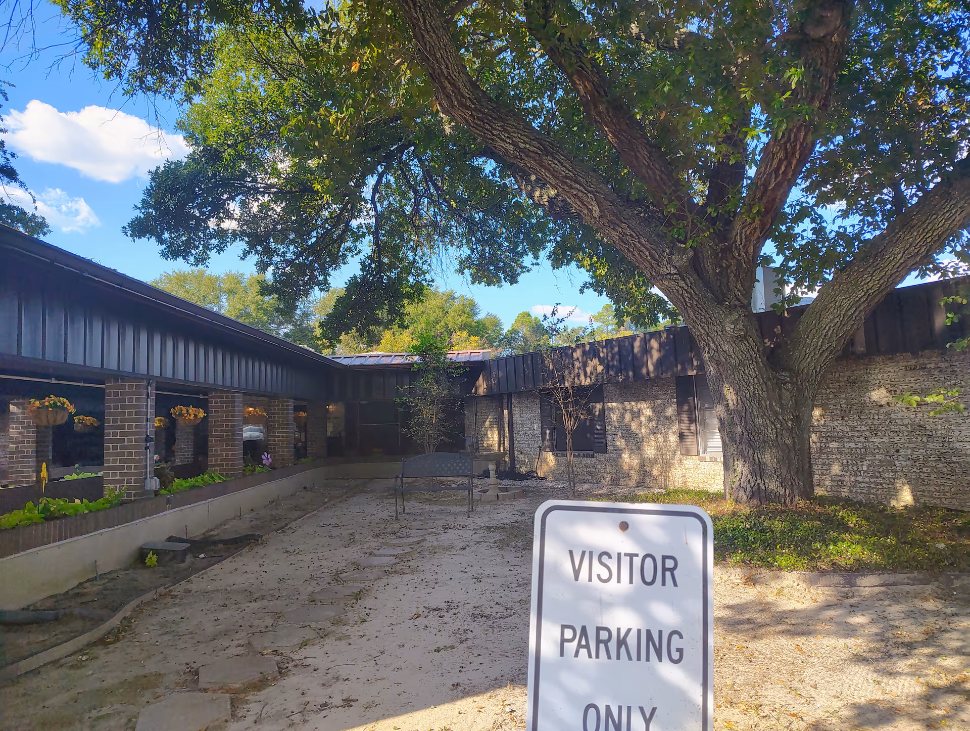 Courtyard with a large tree, brick building, benches, and a visitor parking sign.