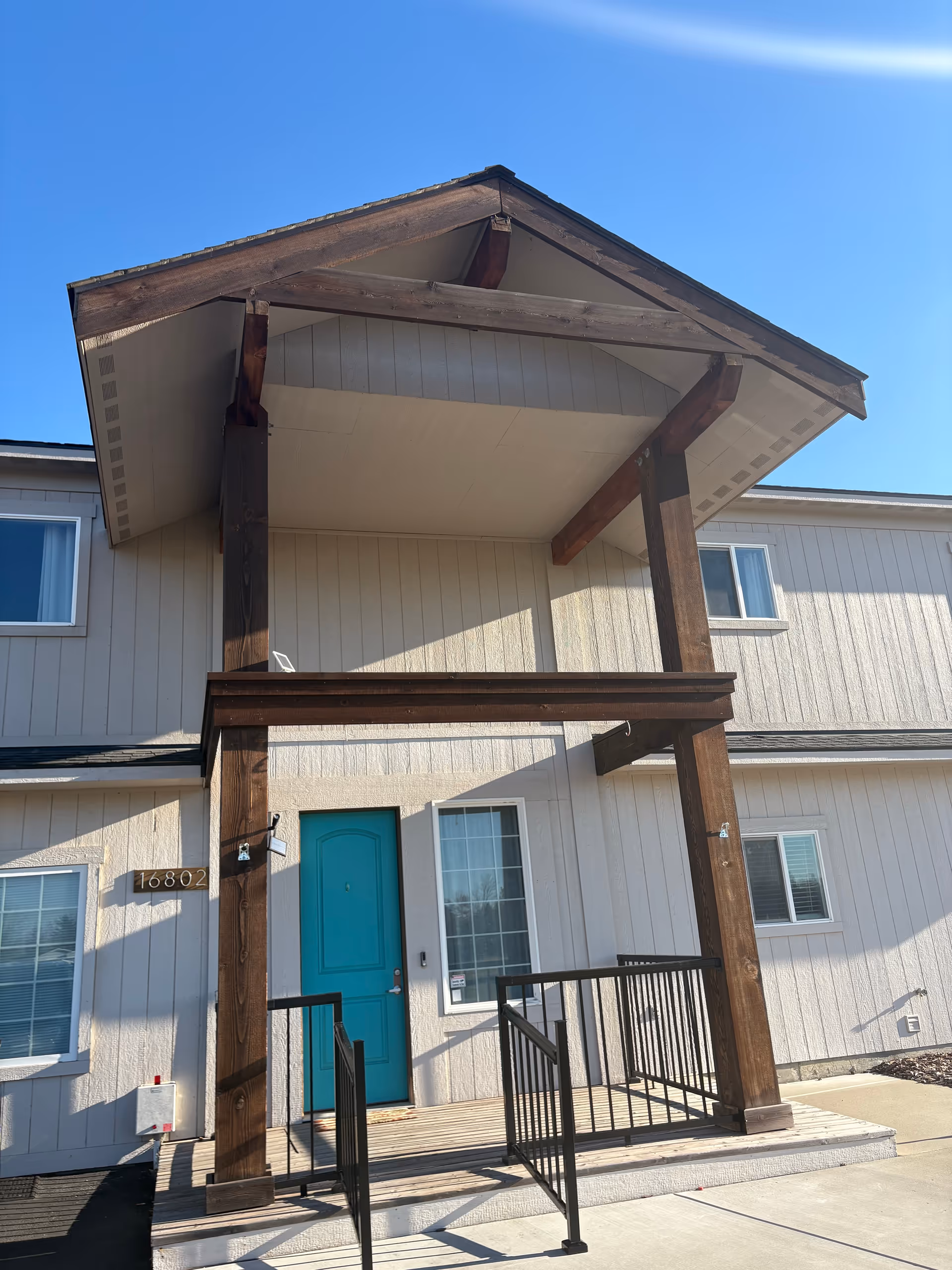Two-story senior living entrance with a teal door and wooden porch pillars under a clear sky.