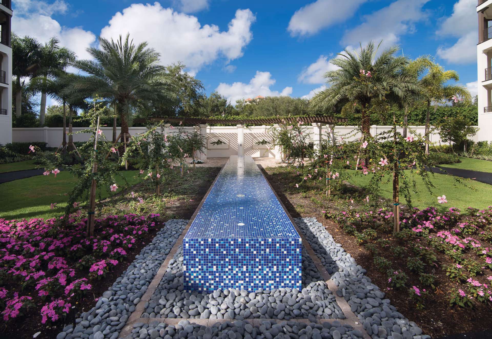 Sunlit courtyard with a long blue tiled water feature flanked by palm trees and flowering plants.