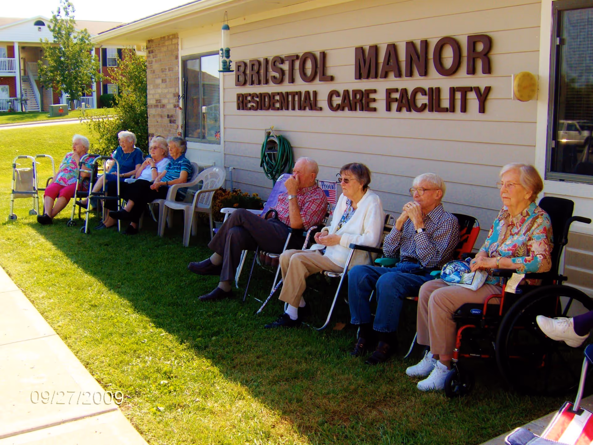 A group of elderly people sitting outside on chairs and wheelchairs along the side of a building with a sign that reads 'Bristol Manor Residential Care Facility'. They are seated on grass near a sidewalk, enjoying the sunny day.