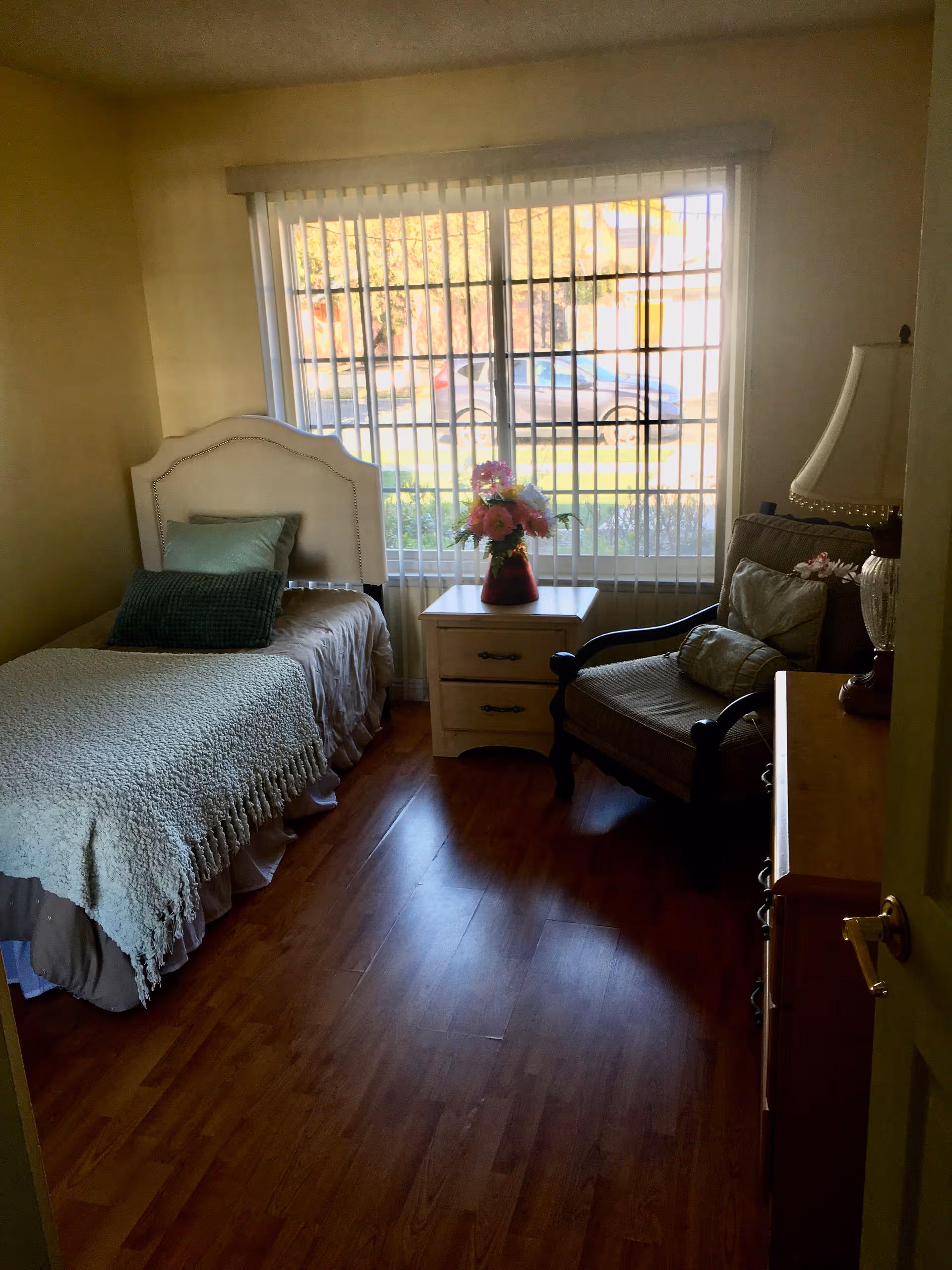 A cozy bedroom with a twin bed, nightstand, chair, dresser, and a large window.