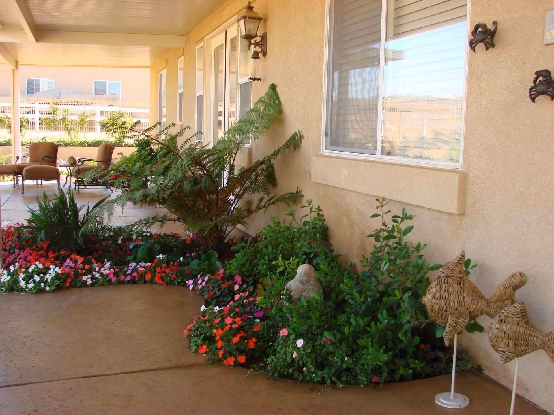 Covered outdoor patio with potted plants, flowers, and seating beside a beige wall.