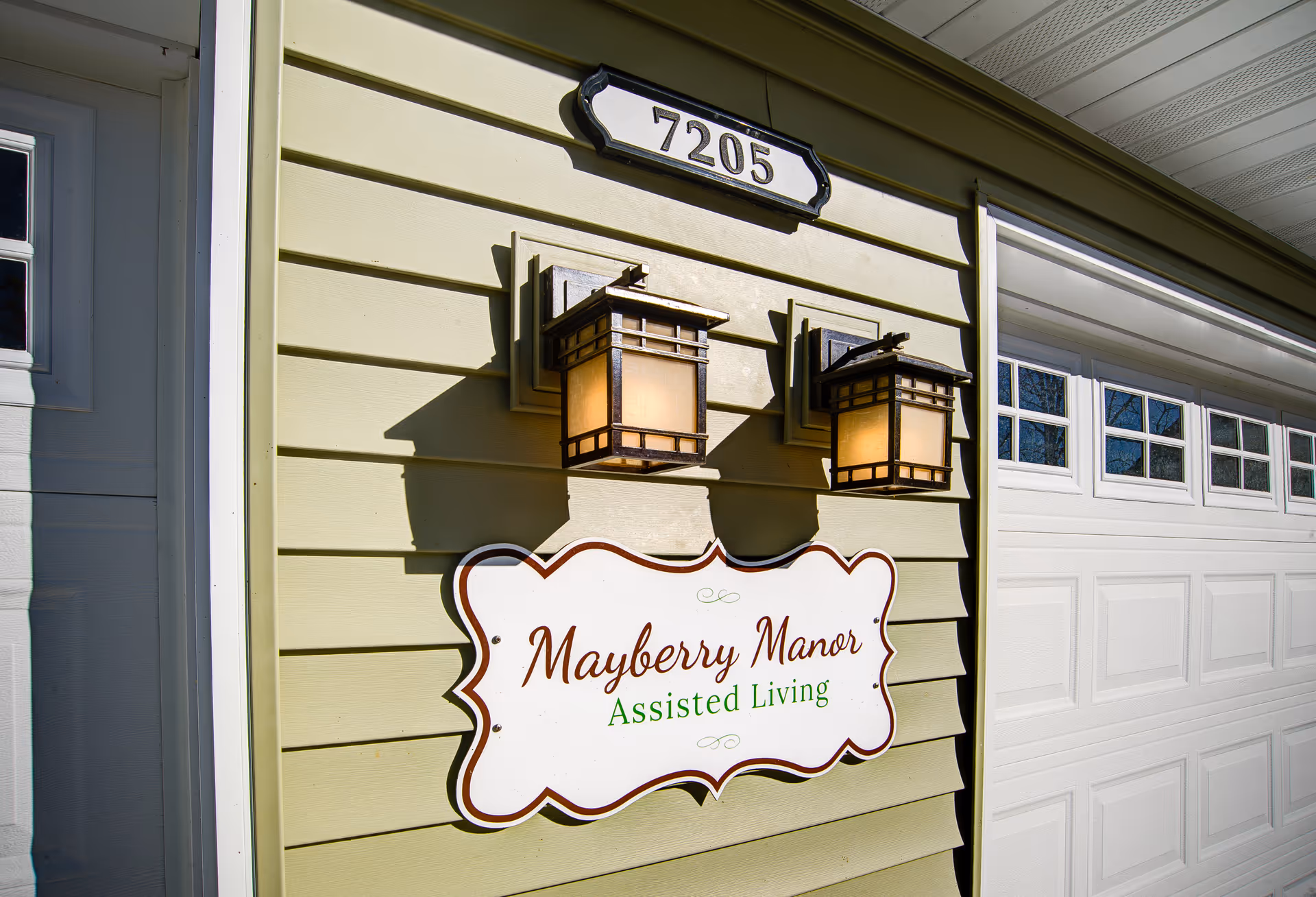 Exterior wall of a building with a sign that reads 'Mayberry Manor Assisted Living' mounted below two decorative outdoor wall lanterns. Above the lanterns is a house number plaque displaying '7205'. Part of a white garage door is visible on the right side.