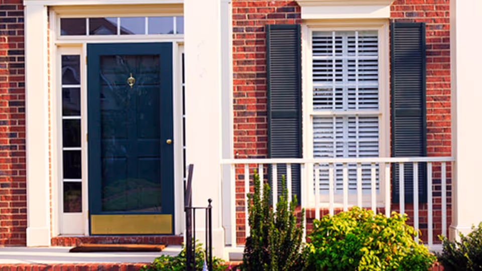 Brick building front with a blue door, white trim, window with shutters, and railing.