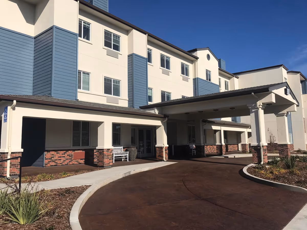 Front entrance and covered drive-up canopy of a multi-story senior living building with benches and landscaping.