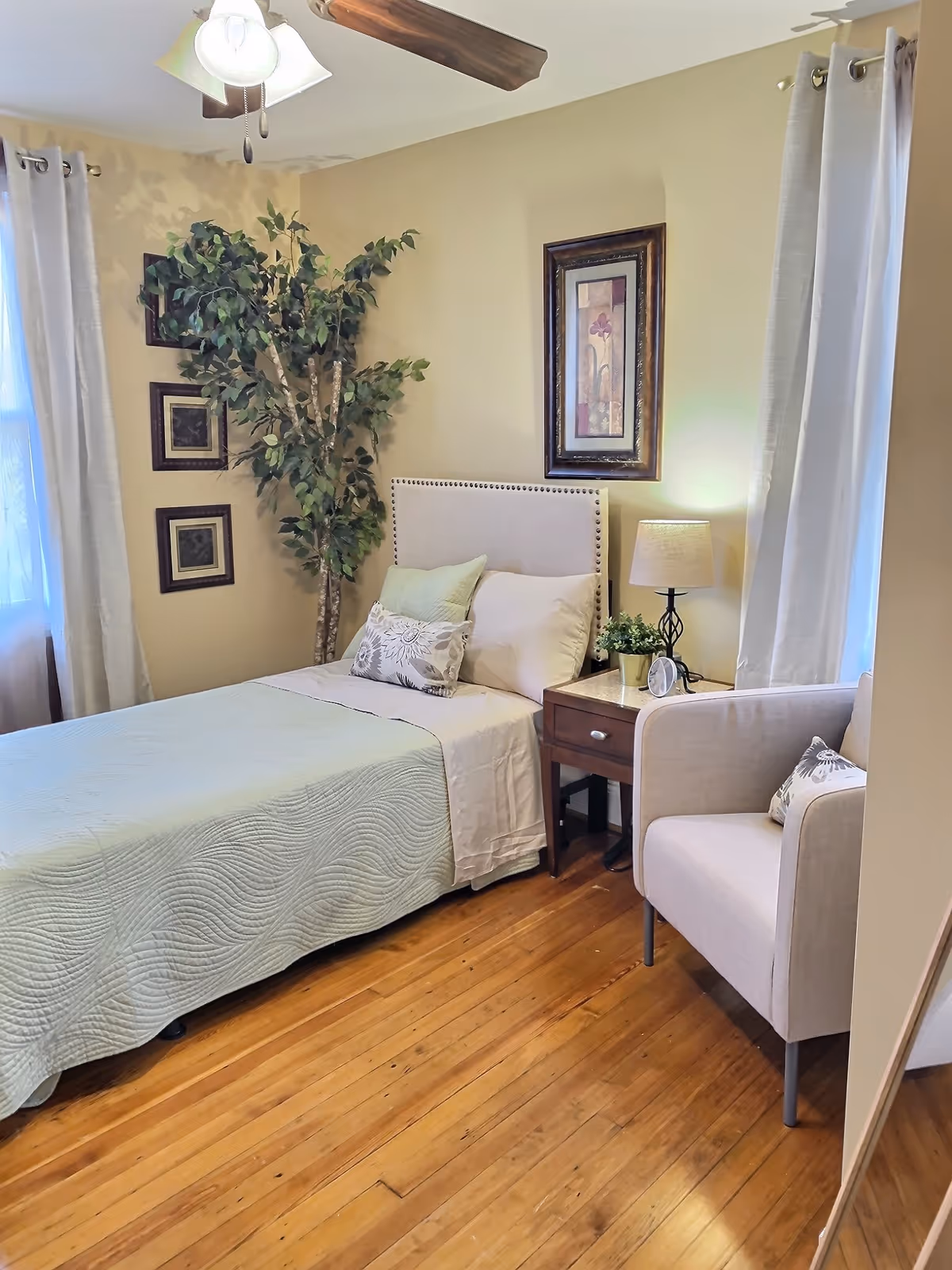 Cozy bedroom in a senior living facility with bed, nightstand, lamp, and chair.