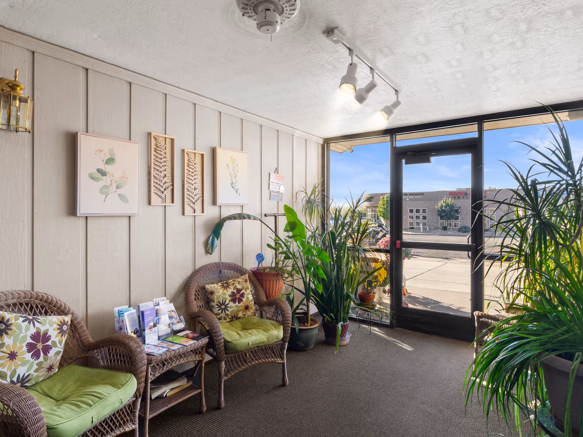 A bright and cozy sitting area with two wicker chairs featuring green cushions and floral pillows, a small wicker table with brochures, several potted plants, and botanical artwork on the wall. A glass door leads outside, letting in natural light.