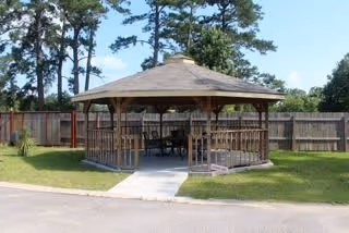 A wooden gazebo with a shingled roof situated on a grassy area, surrounded by a wooden fence and tall trees in the background. There is a paved walkway leading to the gazebo, which contains outdoor furniture inside.