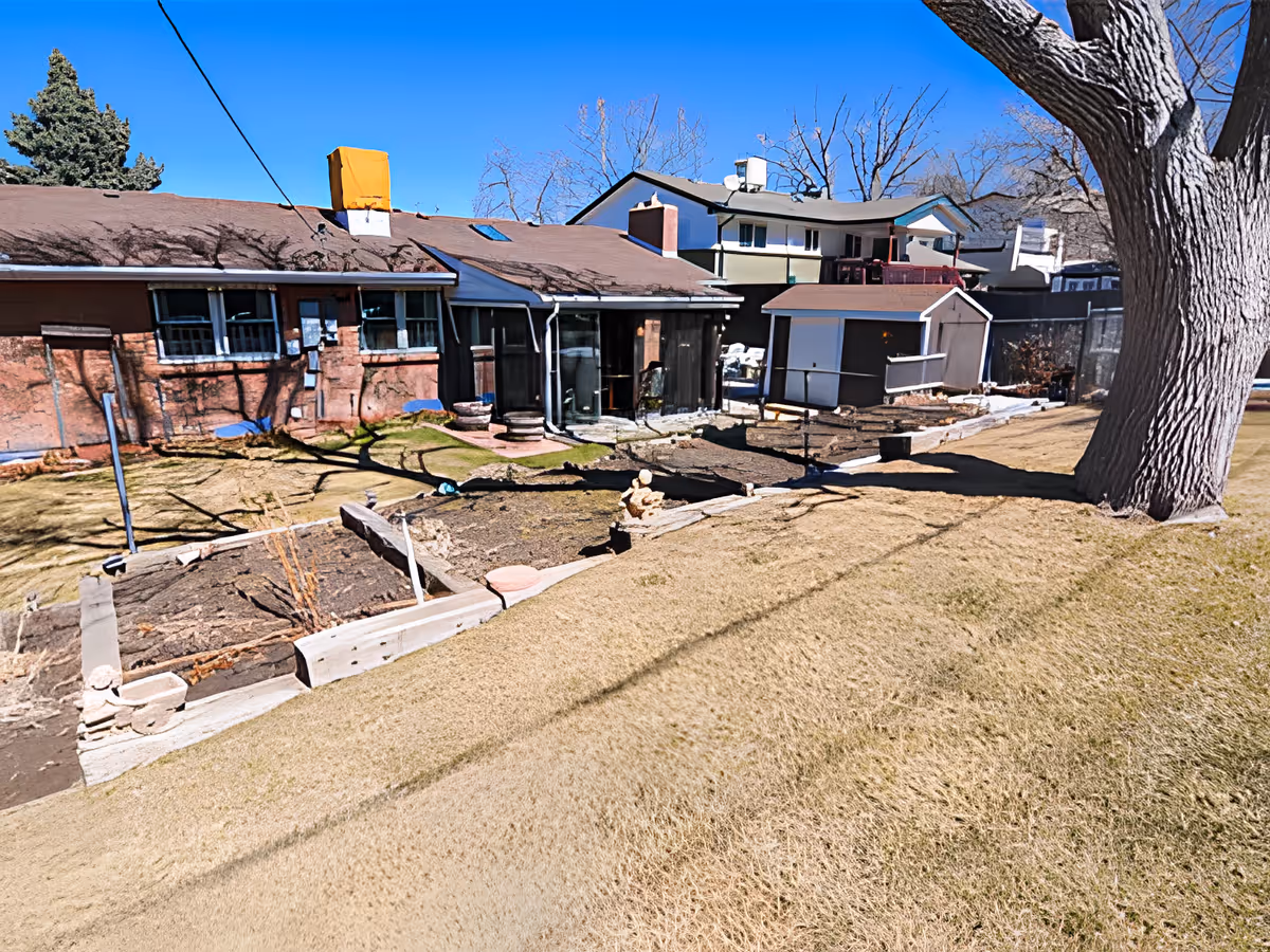 Backyard of a single-story building with raised garden beds, a small shed, patio area and a large tree under a clear sky.