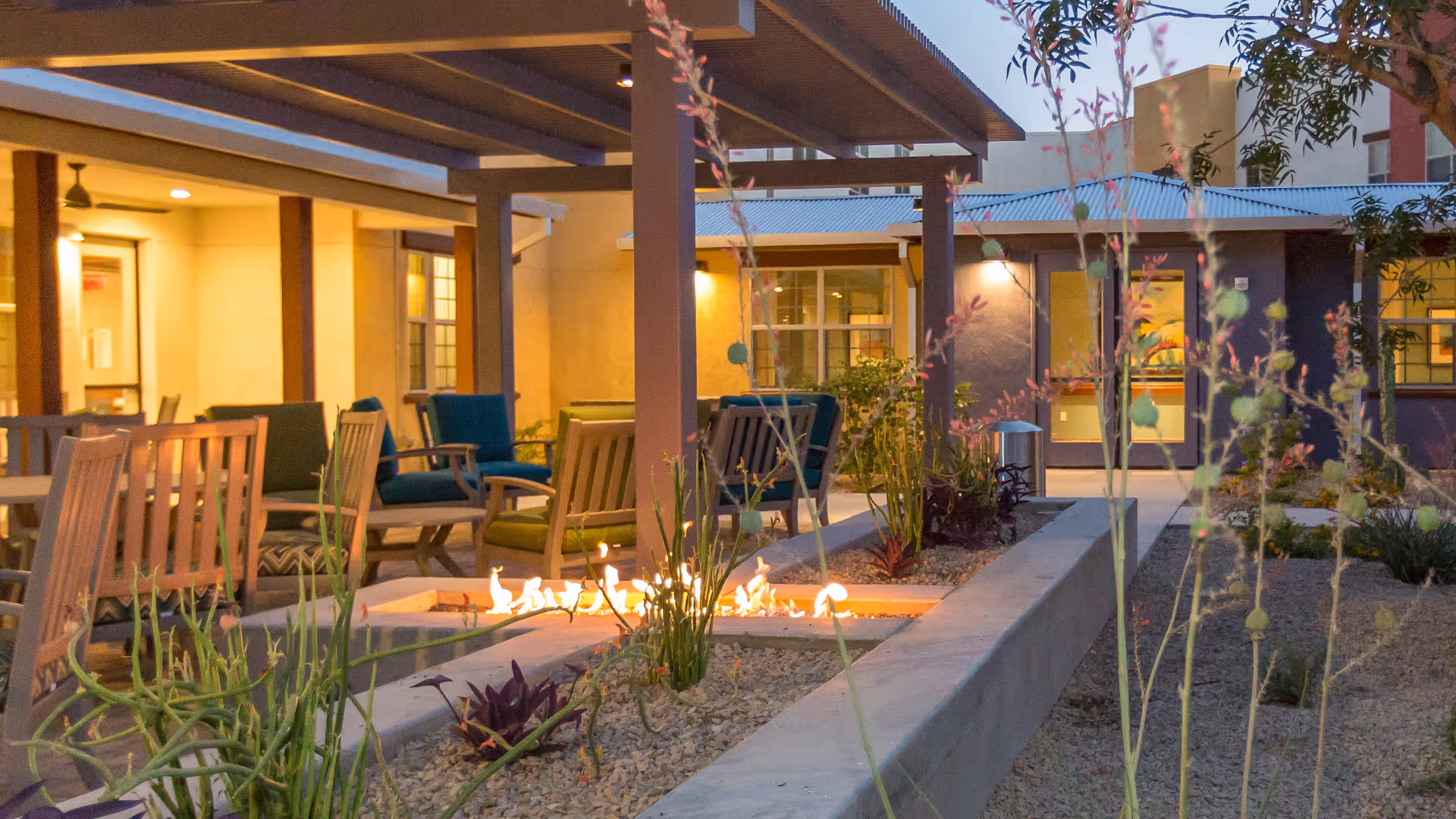 Outdoor seating area at dusk with wooden chairs and cushioned armchairs arranged around a rectangular fire pit. The space is covered by a pergola and surrounded by desert landscaping with gravel and plants. Warm lighting illuminates the building exterior in the background.
