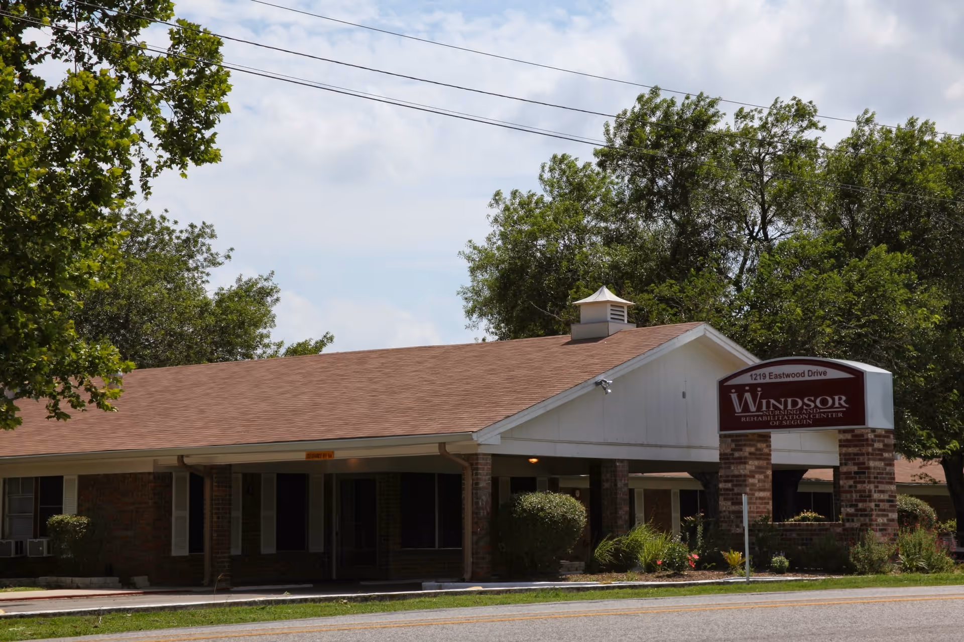 Front exterior of Windsor Nursing and Rehabilitation Center of Seguin showing a low brick building with a covered entrance and the facility sign by the street.