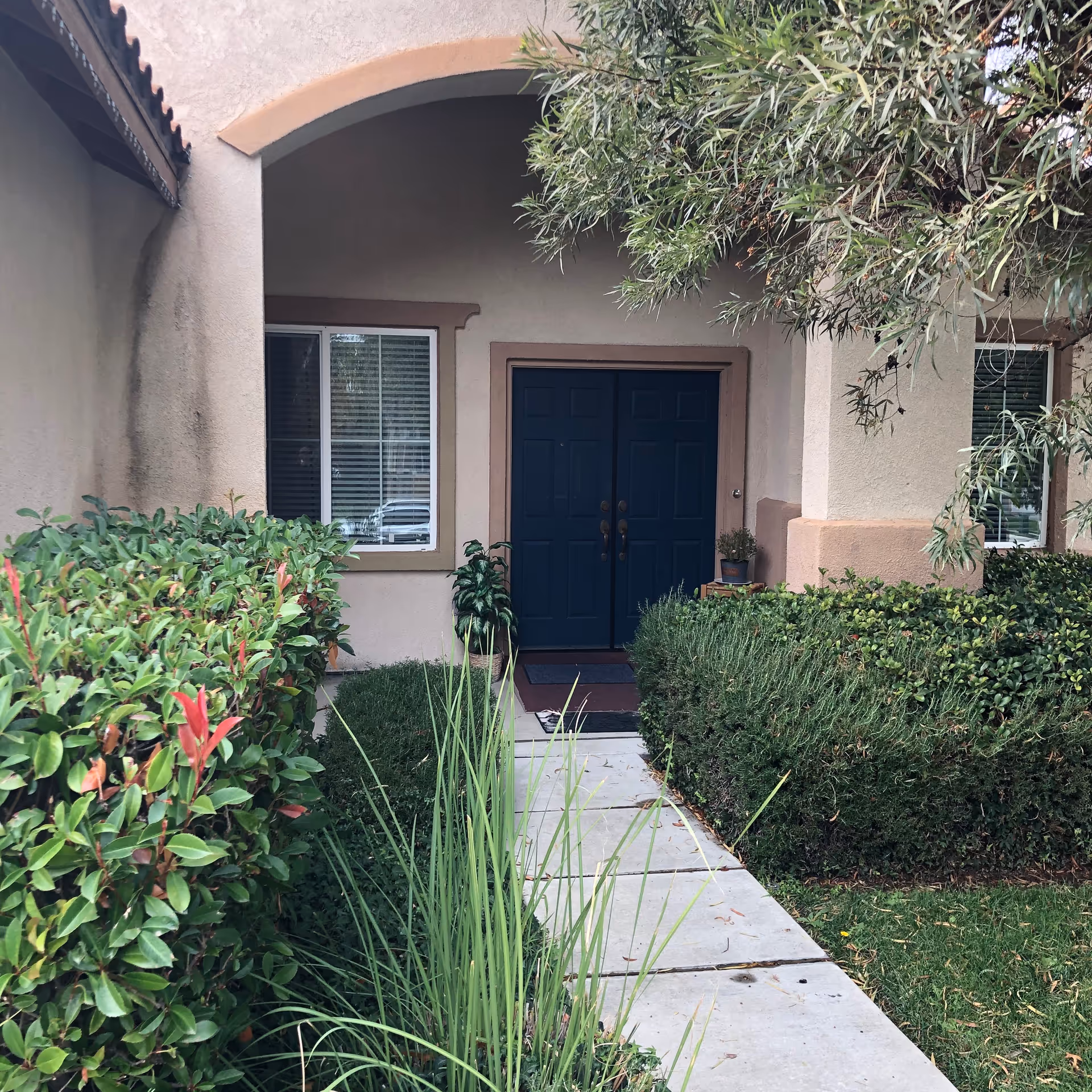A tan stucco senior living facility entrance with a blue double door and surrounding shrubs.