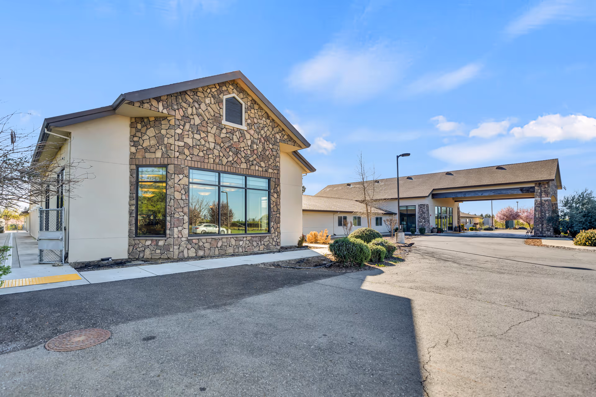 Exterior view of a senior living facility building with stone and beige walls, large windows, and a covered entrance driveway under a clear blue sky.