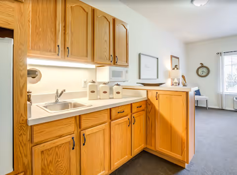 Oak kitchen cabinets, sink, and microwave line a compact kitchenette in a senior living unit.