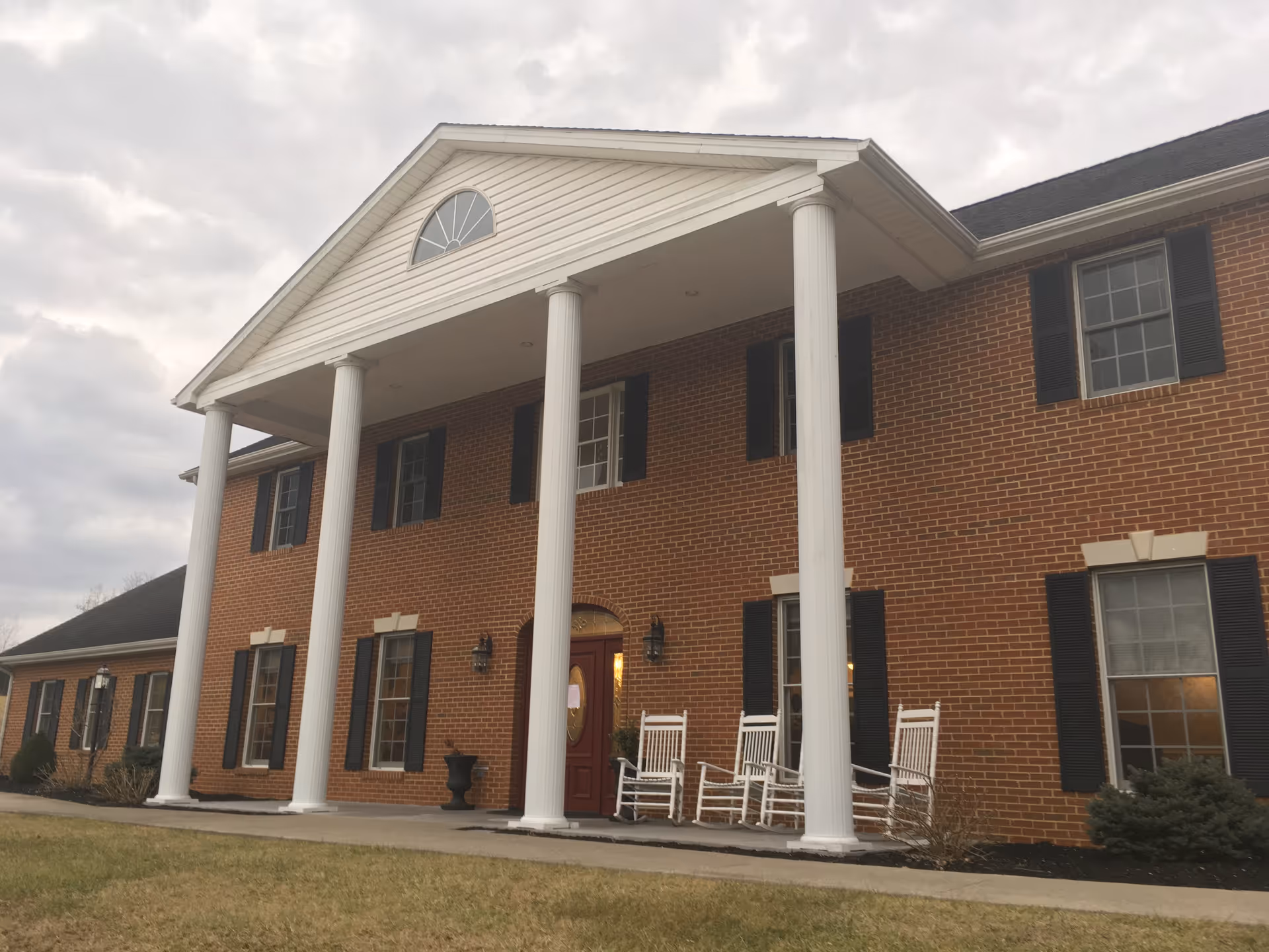 Brick senior living building with a white-columned front porch and rocking chairs.
