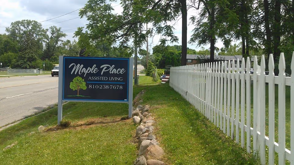 A roadside sign reading "Maple Place Assisted Living" on a grassy lawn beside a white picket fence and trees.