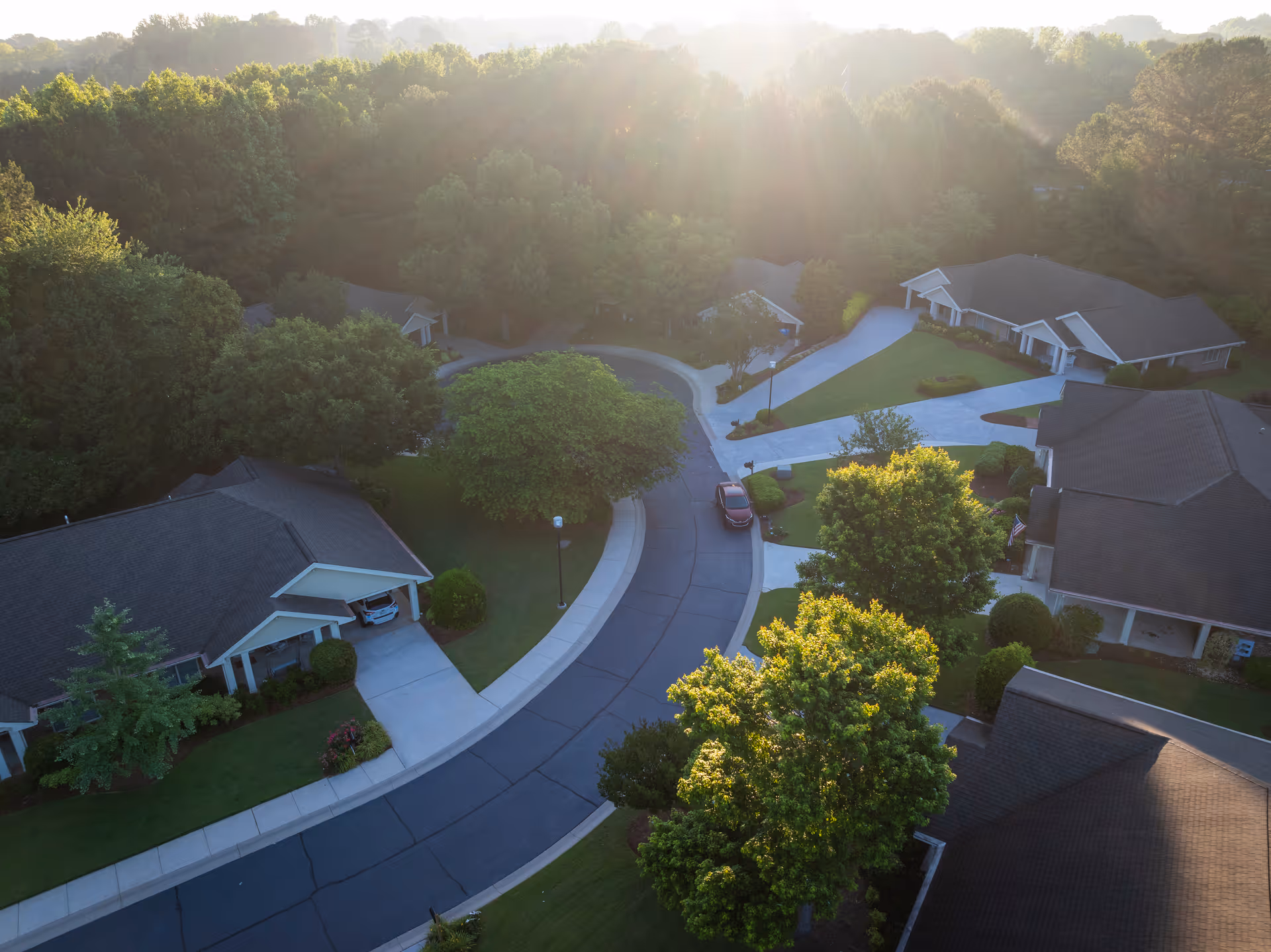 Aerial view of a residential neighborhood within Presbyterian Village, showing several single-story houses with driveways, surrounded by green trees and lawns under soft sunlight.
