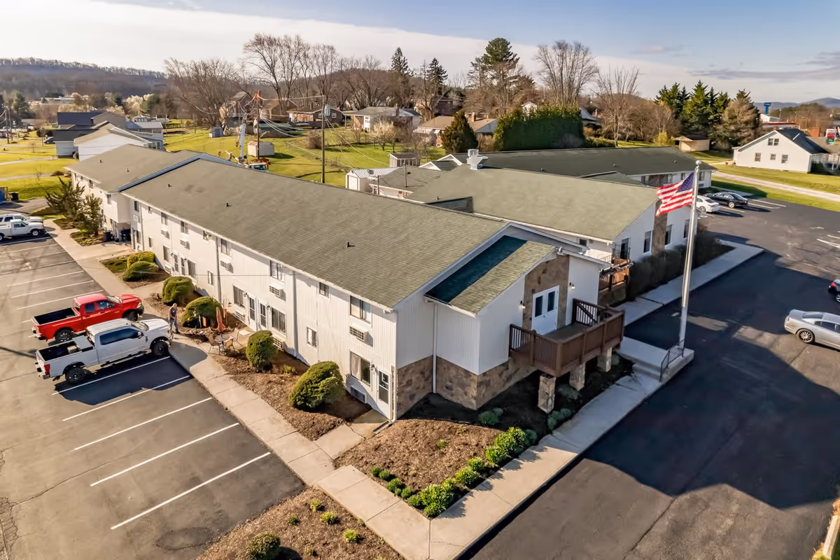 Aerial view of a two-story senior living facility building with a green roof and stone accents, surrounded by parking lots with several parked vehicles. The building has a small balcony and an American flag on a flagpole near the entrance. The surrounding area includes grassy lawns, trees, and neighboring houses under a partly cloudy sky.