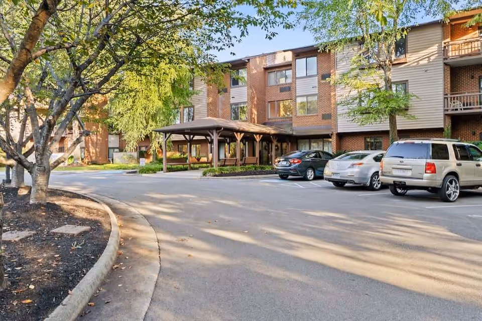 Front exterior of a multi-story senior living building with a covered entrance, parked cars, and trees.
