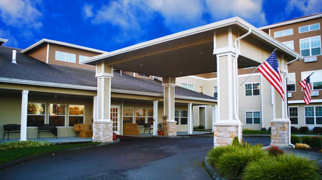 Entrance canopy and front facade of a multi-story senior living building with American flags and a landscaped driveway.