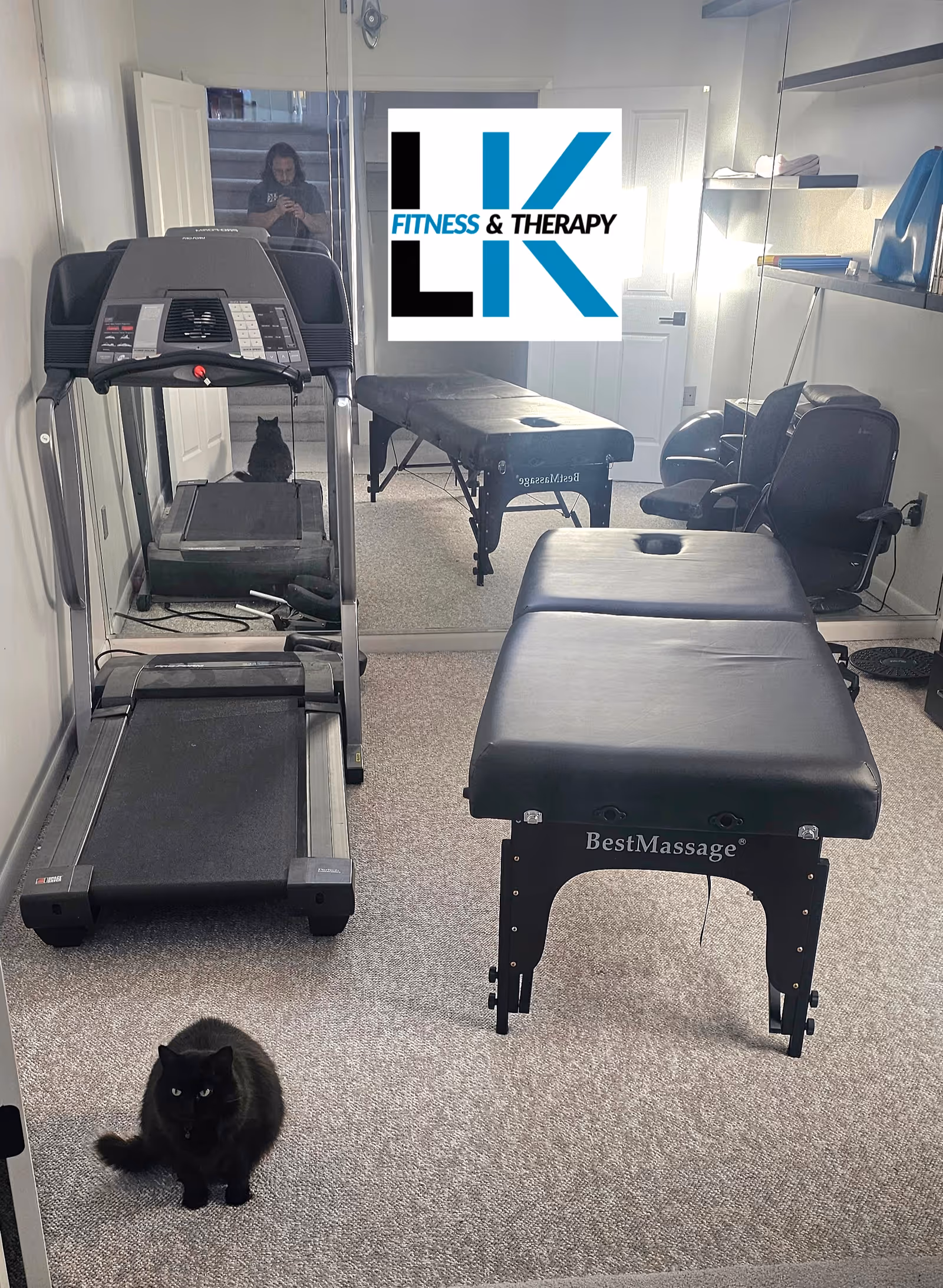 Small interior fitness and therapy room with treadmill, massage table, chairs, and a black cat.