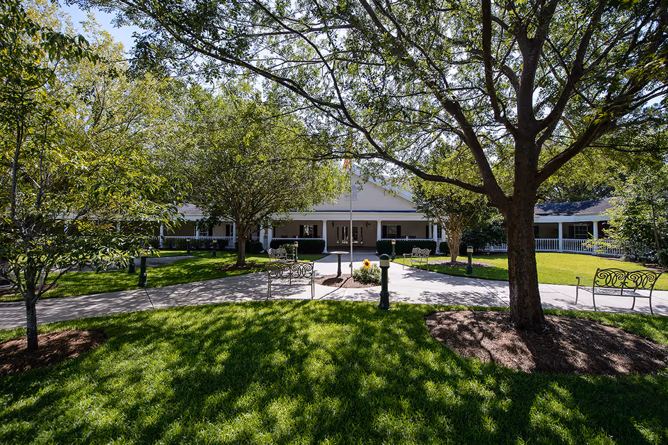 Courtyard and entrance of a senior living facility with a tree-shaded lawn, paved walkways, benches, and a white single-story building with a covered porch.