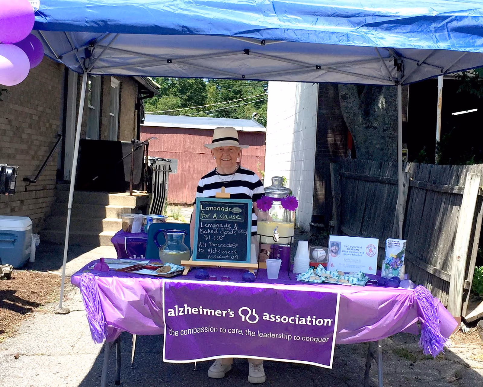 Volunteer stands behind a purple Alzheimer's Association table under a blue outdoor canopy.