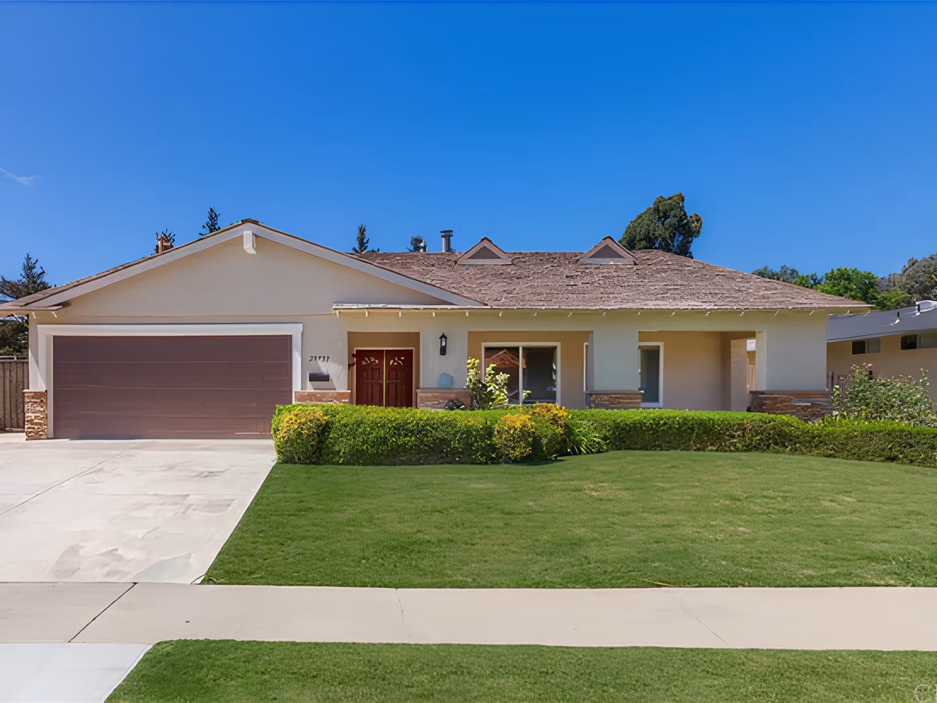 Single-story residential building with a brown garage door, red front door, and well-maintained green lawn and bushes under a clear blue sky.