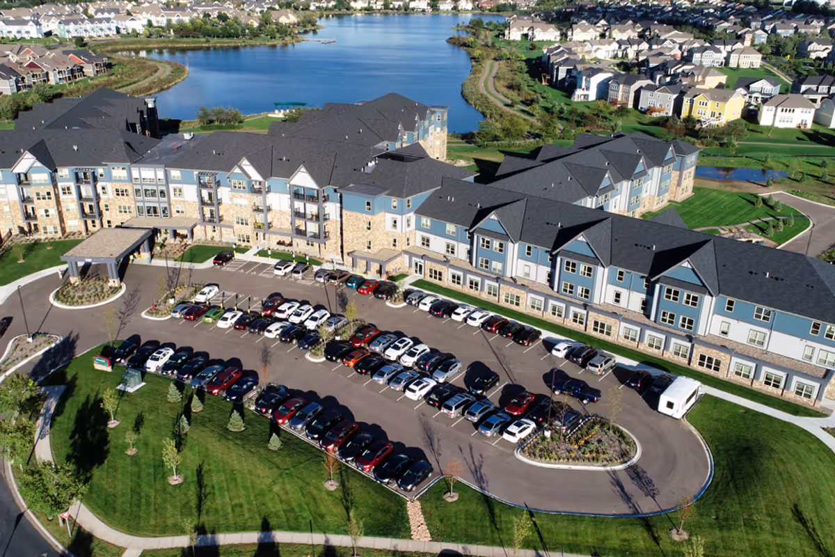Aerial view of Orchard Path senior living facility showing a large multi-story building with blue and beige exterior, surrounded by a parking lot filled with cars, green lawns, trees, and a lake in the background with residential houses on the far side.