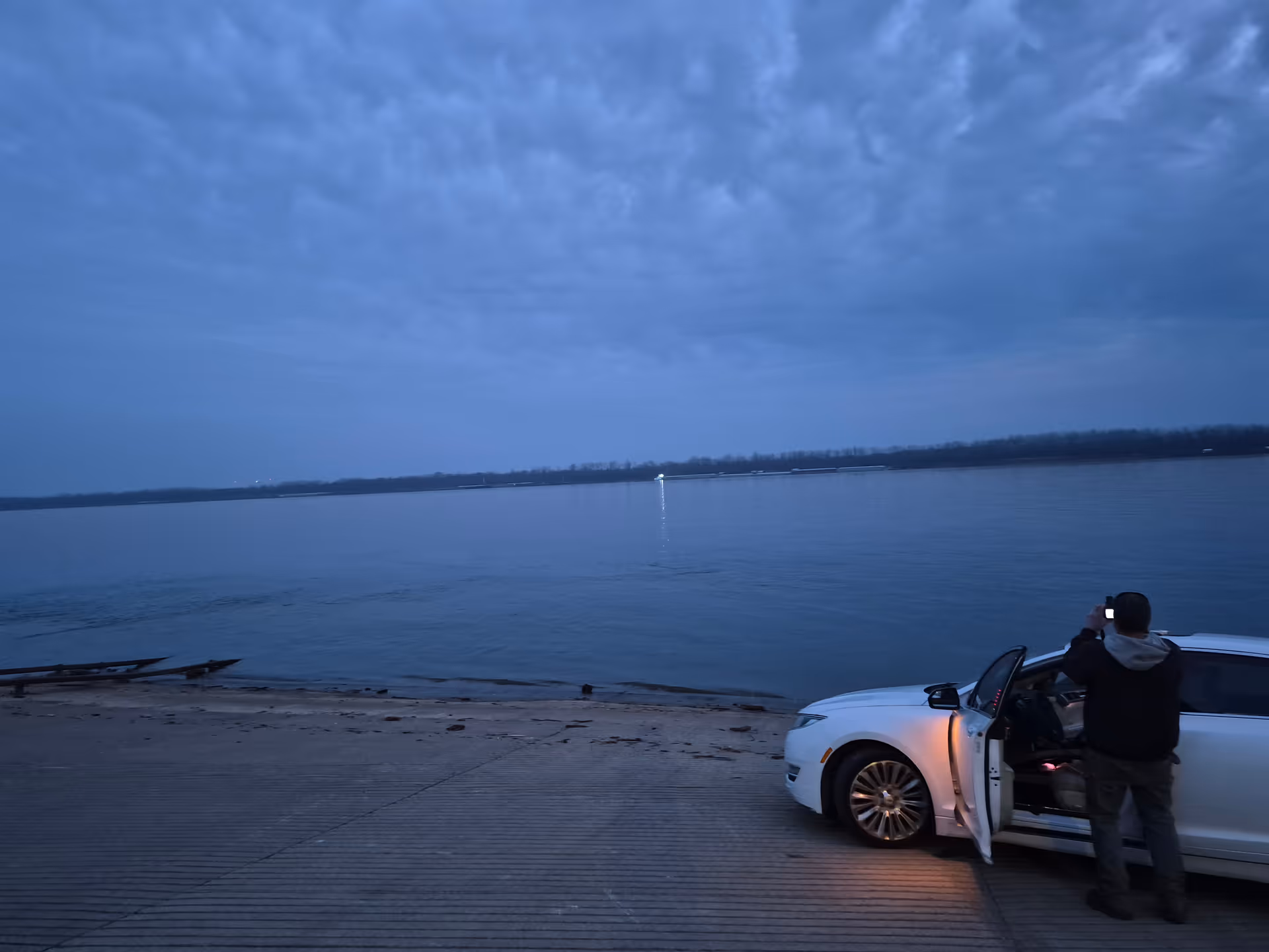 A man stands beside an open white car door on a waterfront promenade.