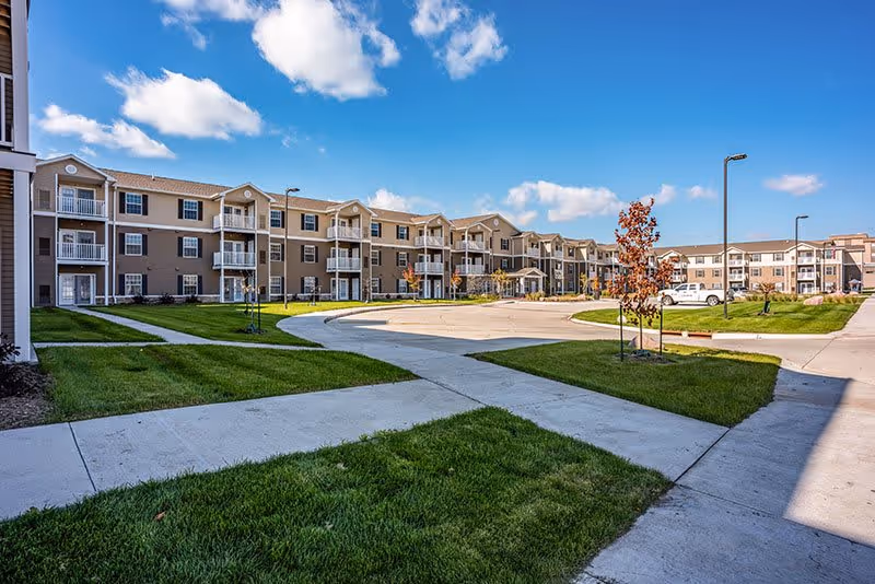 Exterior view of a senior living facility with multiple three-story buildings featuring balconies, surrounded by well-maintained green lawns, sidewalks, and a circular driveway under a partly cloudy blue sky.