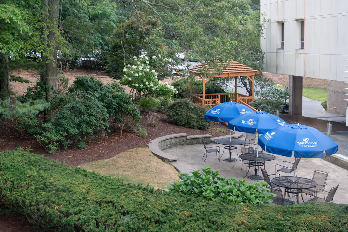 Outdoor patio area with several round metal tables and chairs, each table shaded by a blue umbrella with the Water's Edge Rehab and Nursing Center at Port Jefferson logo. The patio is surrounded by greenery, including bushes, trees, and a wooden gazebo in the background. Part of a building is visible on the right side.