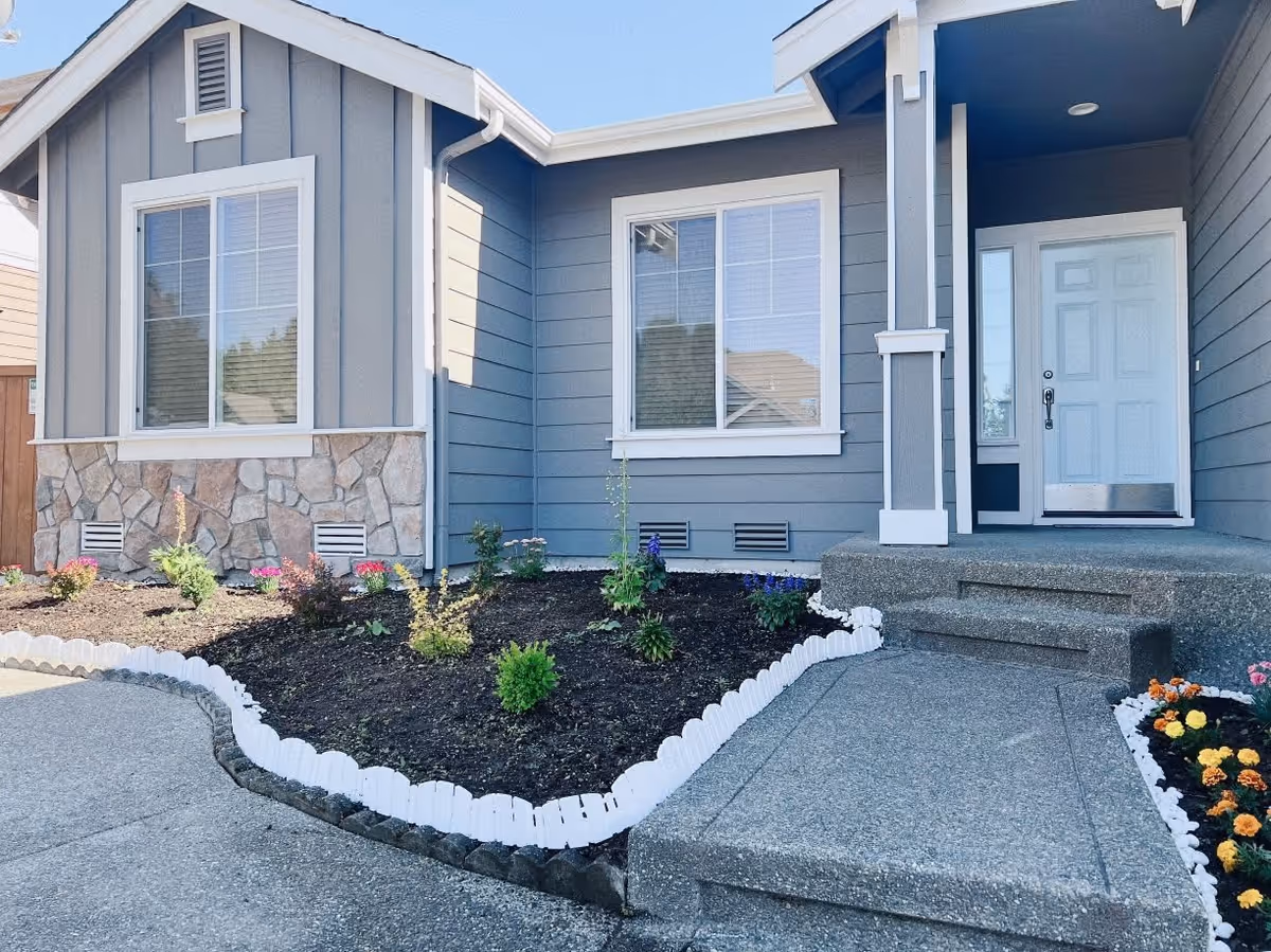 Front entrance of a gray single-story home with a concrete walkway, small landscaped garden beds, and white-trimmed windows and door.